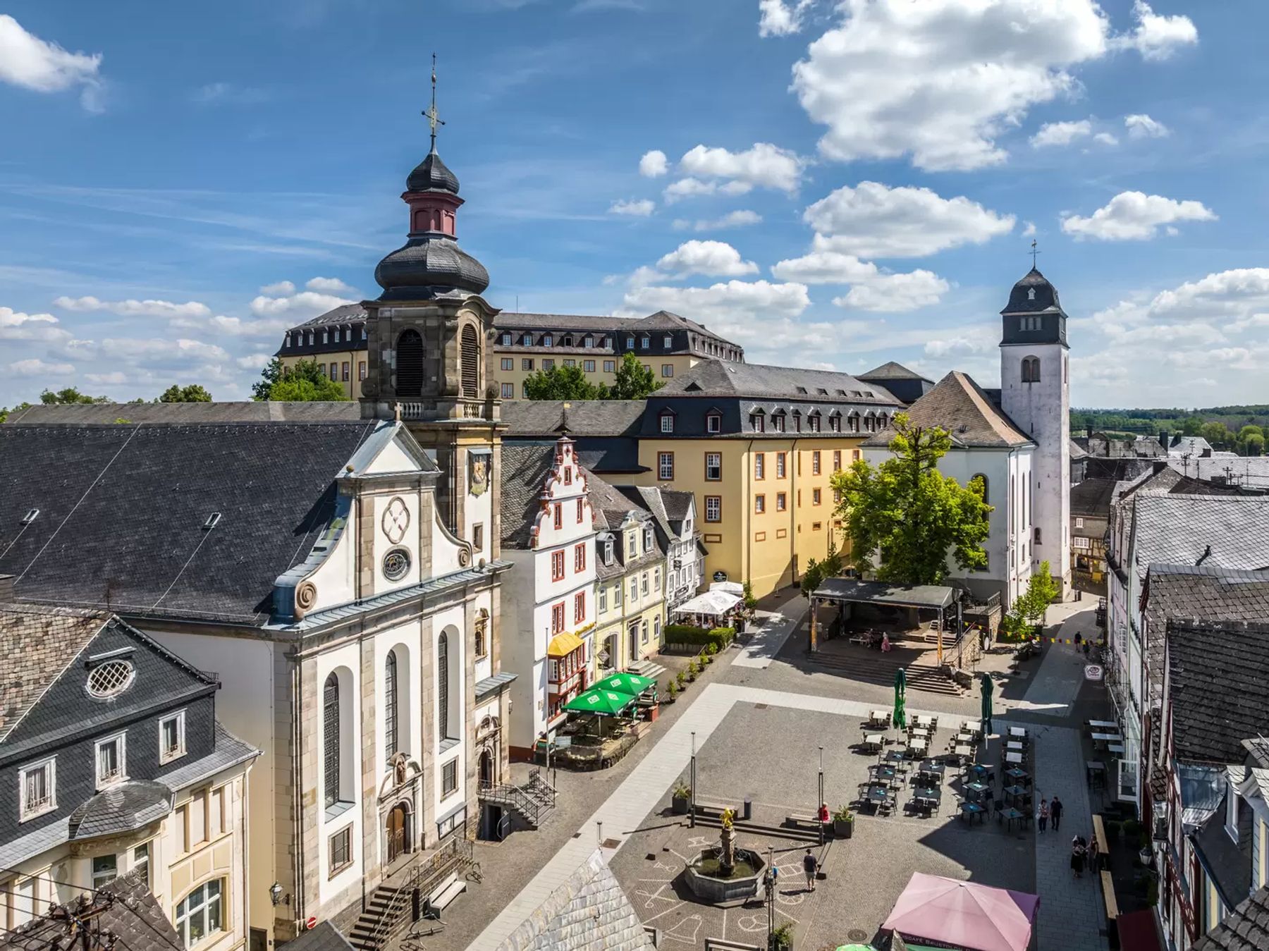 Ein malerischer europäischer Stadtplatz mit historischen Gebäuden, Sitzgelegenheiten im Freien, einem Uhrenturm und Menschen, die den sonnigen Tag unter einem blauen Himmel mit vereinzelten Wolken genießen.