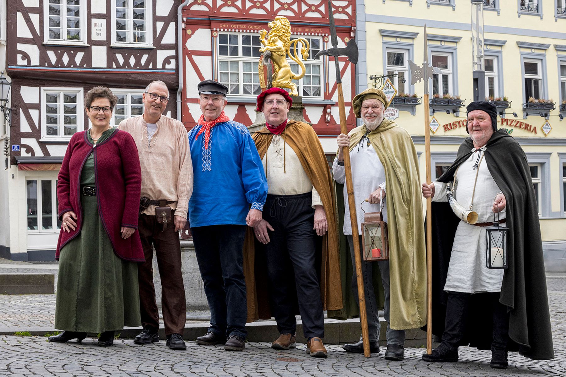 Sechs Erwachsene in historischen Kostümen stehen lächelnd vor Fachwerkhäusern und einem Brunnen auf einem kopfsteingepflasterten Stadtplatz; einige halten Laternen, Stäbe und tragen Umhänge oder Hüte.