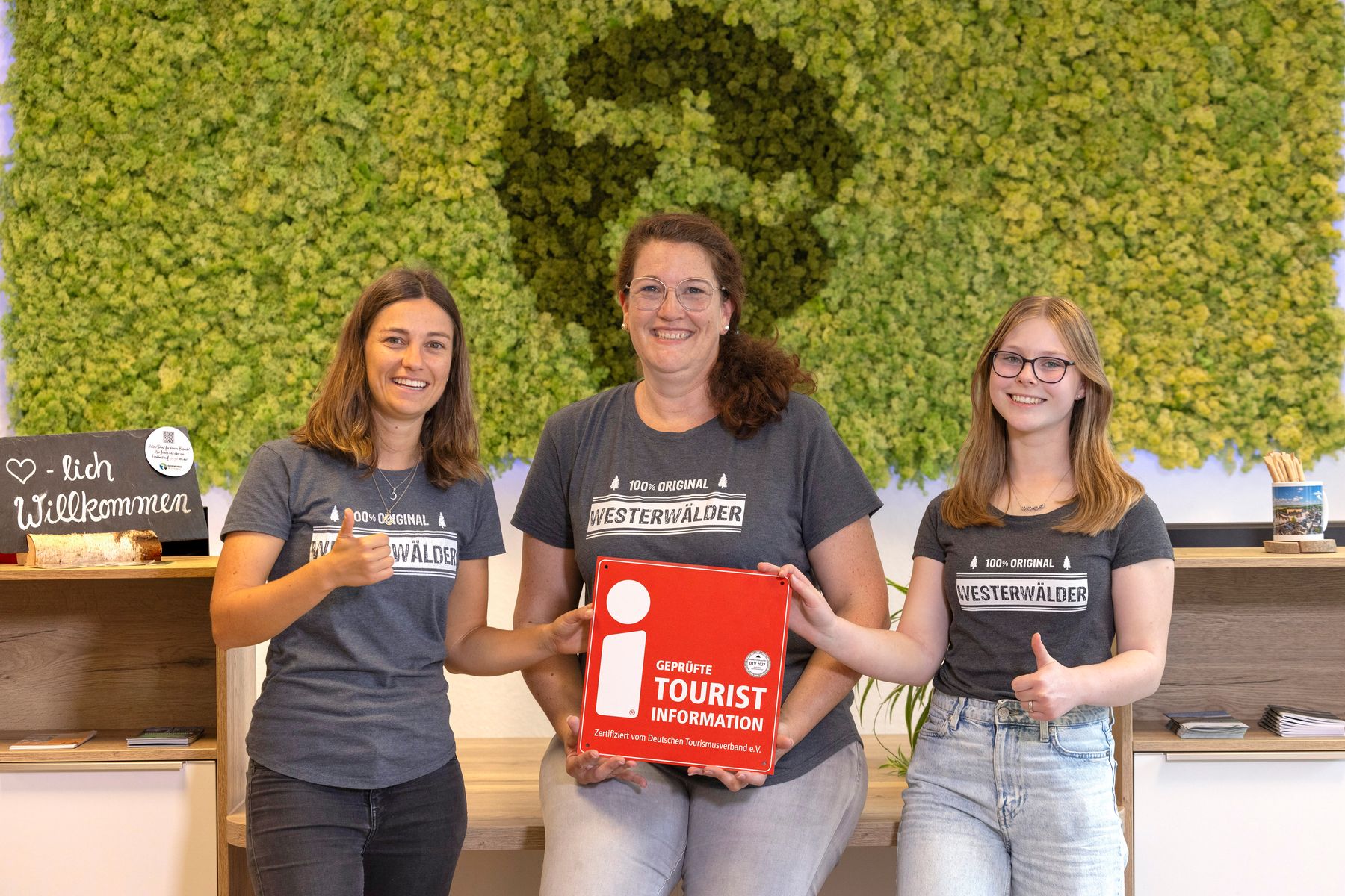 Drei lächelnde Frauen in passenden "Westerwälder"-Shirts stehen drinnen und zeigen die Daumen nach oben. Die Frau in der Mitte hält eine rote Schachtel mit der Aufschrift "Tourist Information". Auf der linken Seite steht ein herzförmiges Willkommen-Schild.