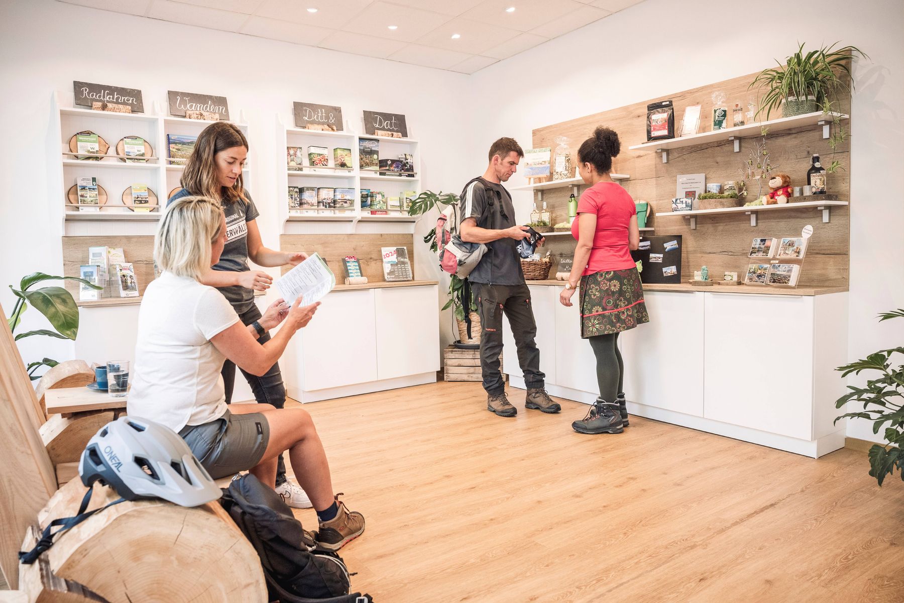 Vier Personen in legerer Outdoor-Kleidung unterhalten sich in einem hellen, modernen Laden mit Holzfußboden und Regalen mit Lebensmitteln und lokalen Produkten. Zwei unterhalten sich an der Theke, während zwei andere sitzen und in Papieren blättern.