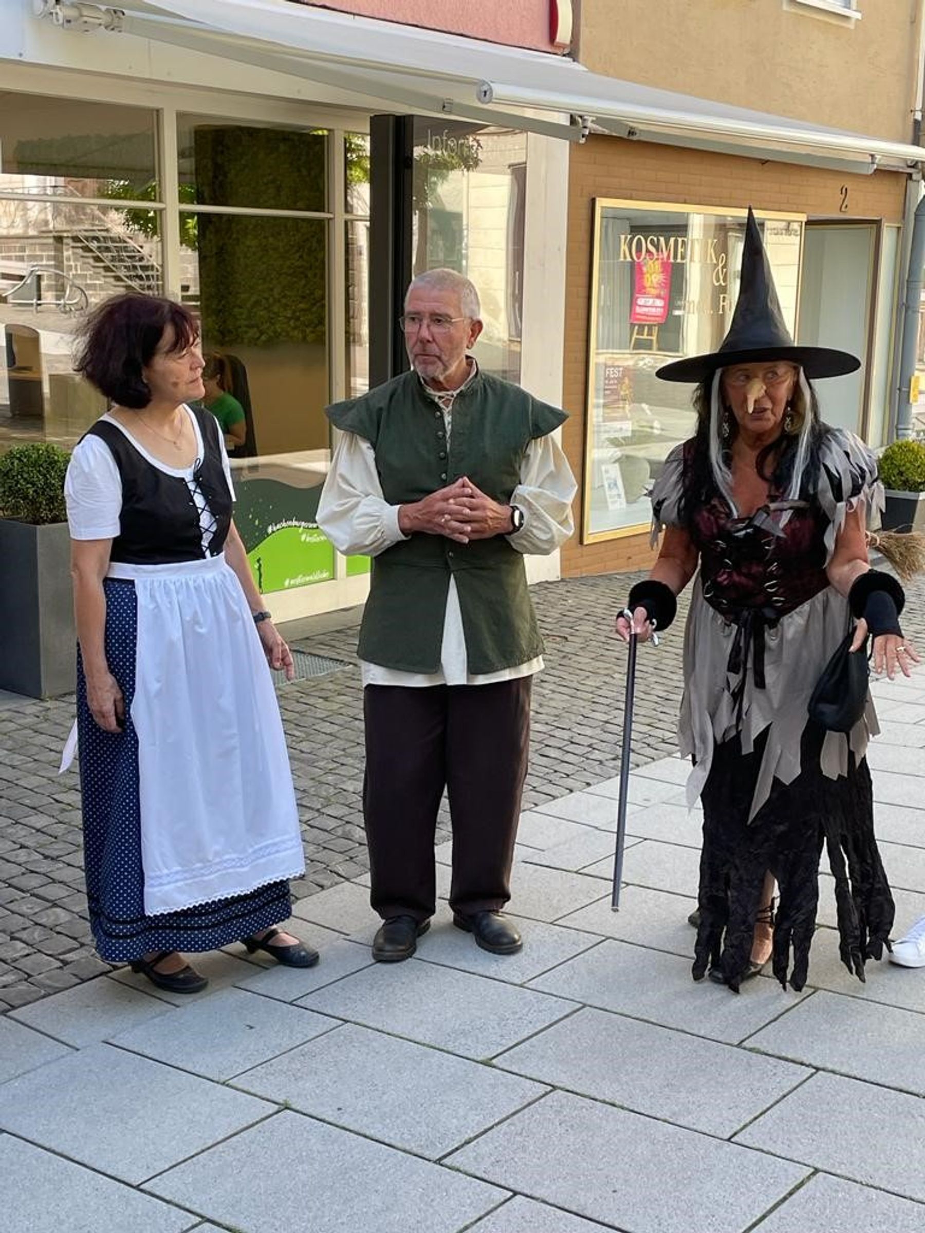 Drei Erwachsene in Kostümen stehen auf einem städtischen Bürgersteig; eine Frau trägt ein Dienstmädchenkostüm, ein Mann ist mittelalterlich gekleidet, und eine weitere Frau trägt ein Hexenkostüm mit Hut und Stab.
