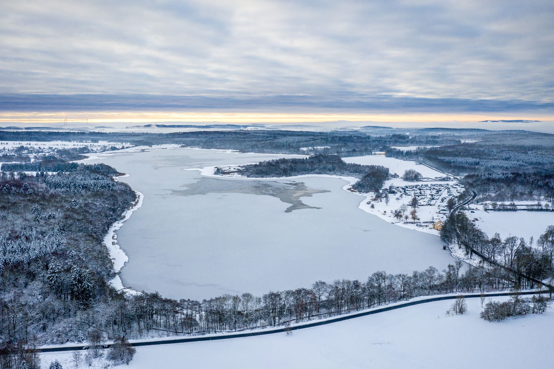 Luftaufnahme eines großen zugefrorenen Sees, umgeben von schneebedeckten Bäumen und Feldern unter einem bewölkten Himmel, mit einer Straße, die sich am Rande des Sees entlang schlängelt, und einem fernen Horizont.