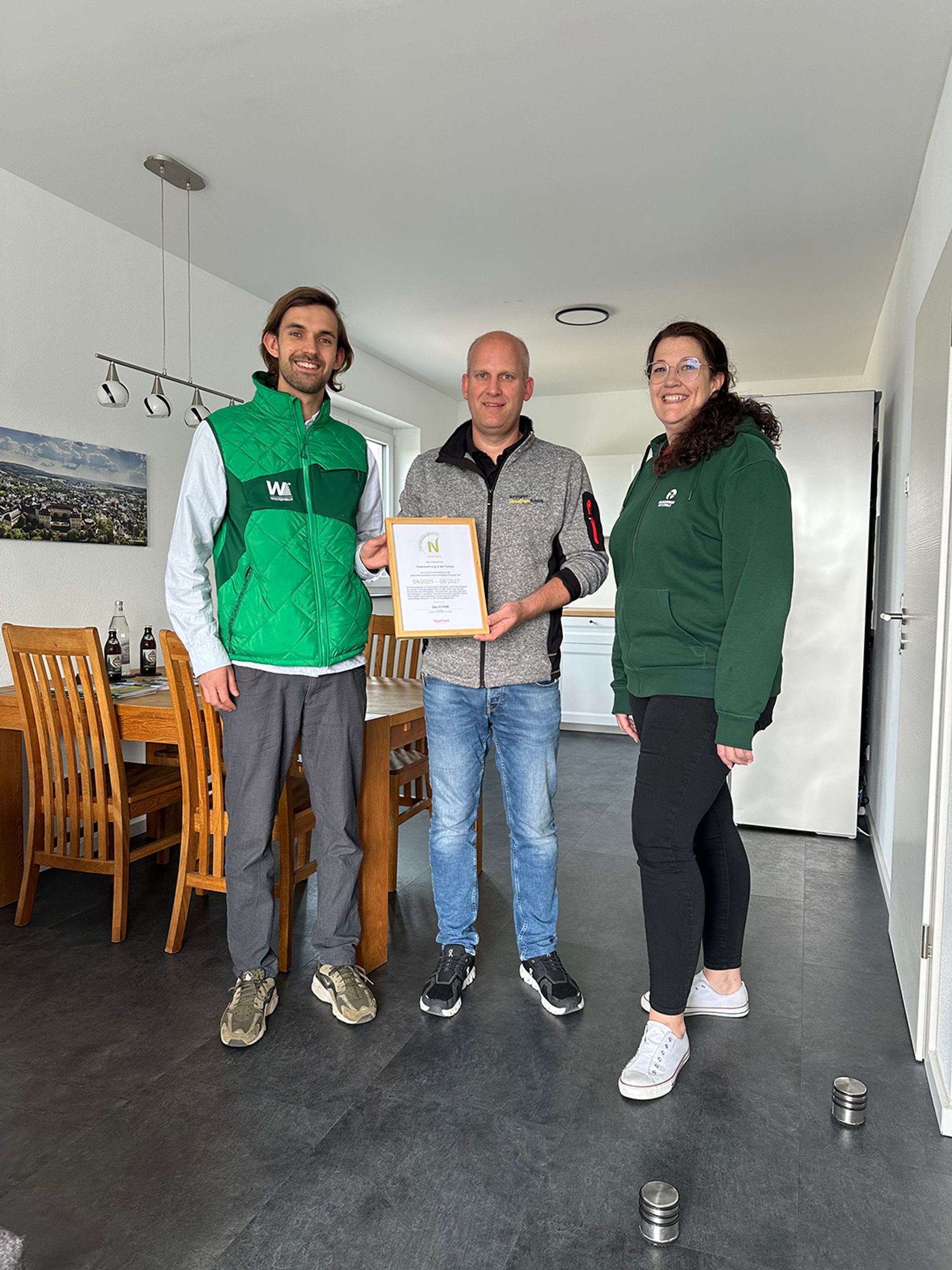 Drei Personen stehen in einem Haus und lächeln in die Kamera. Der Mann in der Mitte hält eine gerahmte Urkunde in der Hand. Sie sind leger gekleidet, und im Hintergrund ist ein hölzerner Esstisch mit Stühlen zu sehen.