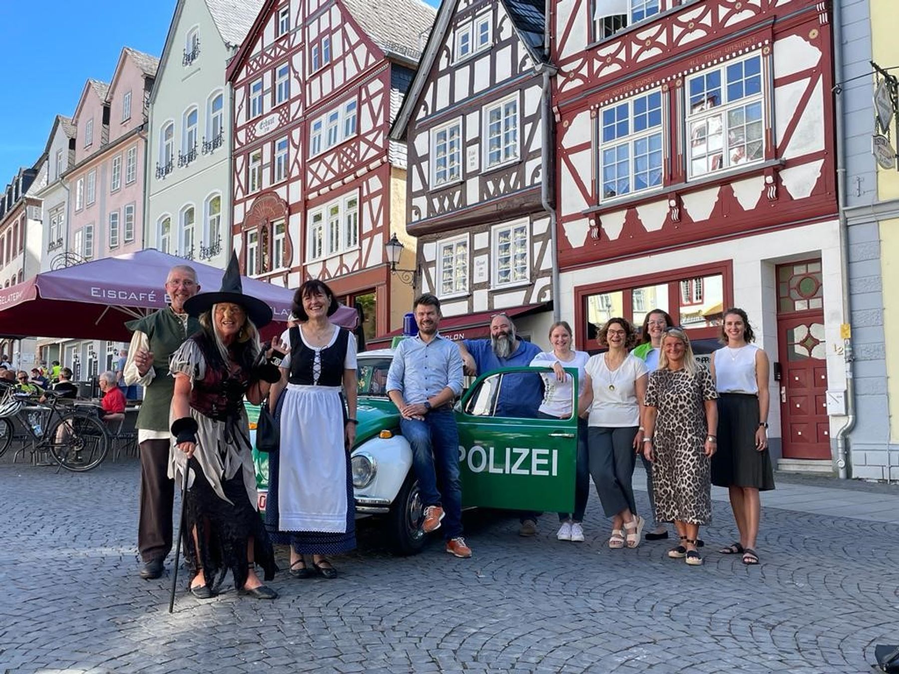 Eine Gruppe von Menschen, einige in Kostümen, andere in Freizeitkleidung, steht lächelnd vor einem alten grünen Polizeiauto auf einer Kopfsteinpflasterstraße, die von bunten, traditionellen deutschen Gebäuden gesäumt ist.