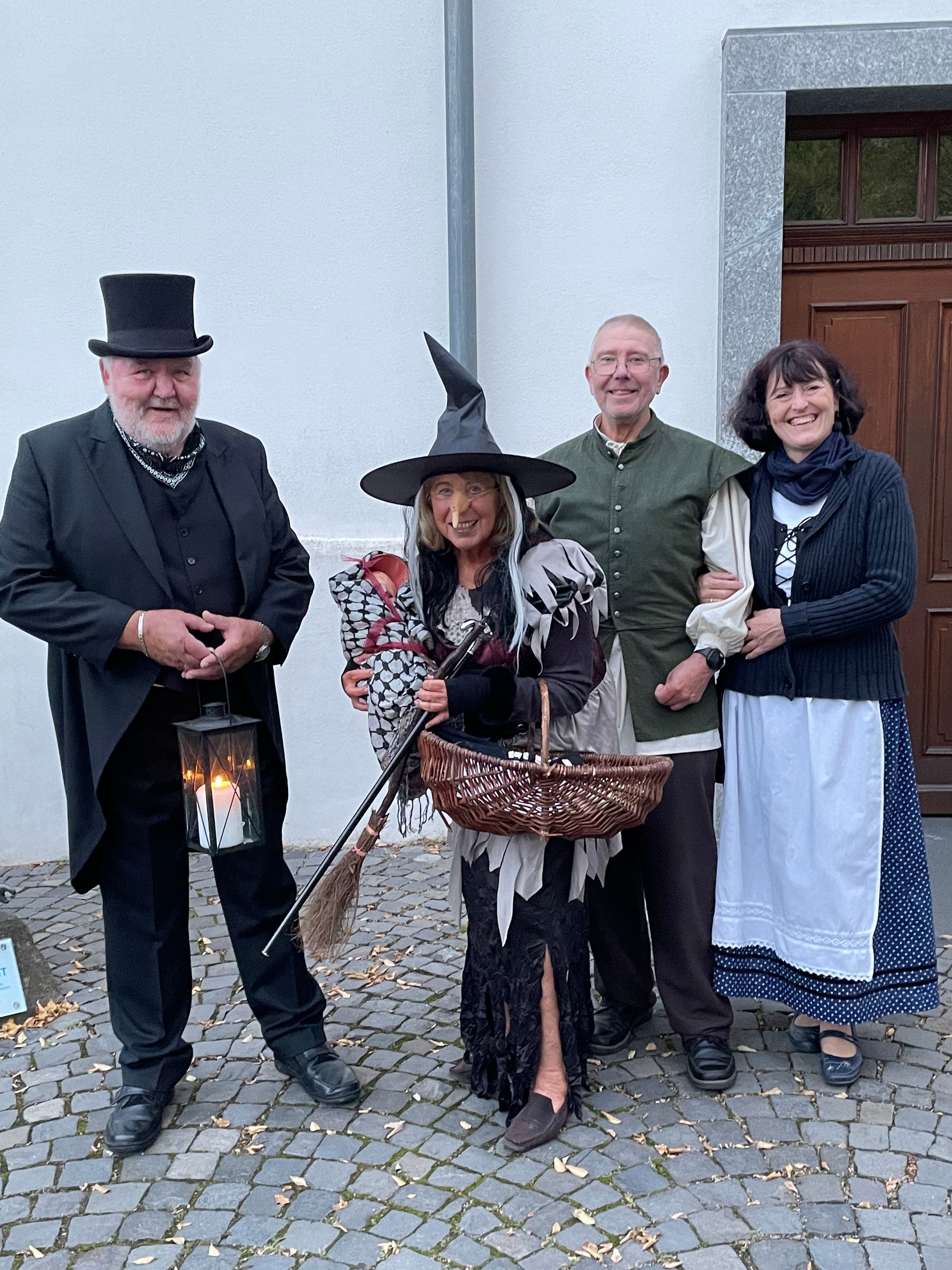 Vier Erwachsene in Kostümen stehen auf einem Kopfsteinpflasterweg. Einer trägt einen Zylinder und hat eine Laterne in der Hand, ein anderer ist als Hexe mit Besen und Korb gekleidet, während die anderen traditionelle Kleidung tragen und in die Kamera lächeln.