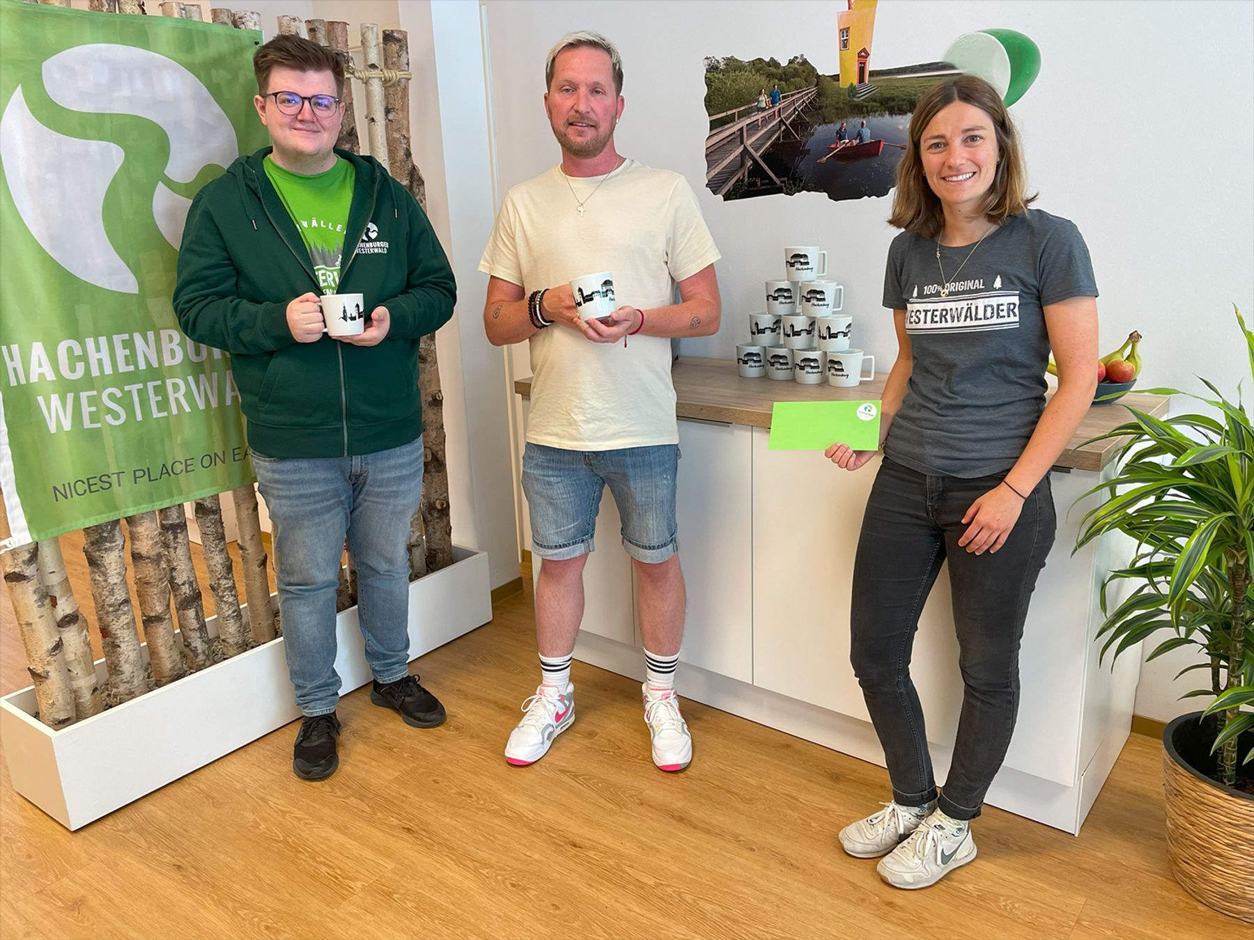 Drei Personen stehen in einem Haus und lächeln in die Kamera. Zwei halten Tassen und eine eine Karte. Eine Pyramide von Tassen ist auf einem Tresen neben ihnen gestapelt. Auf einem Schild steht "WESTERWALD". Die Umgebung wirkt freundlich und ungezwungen.