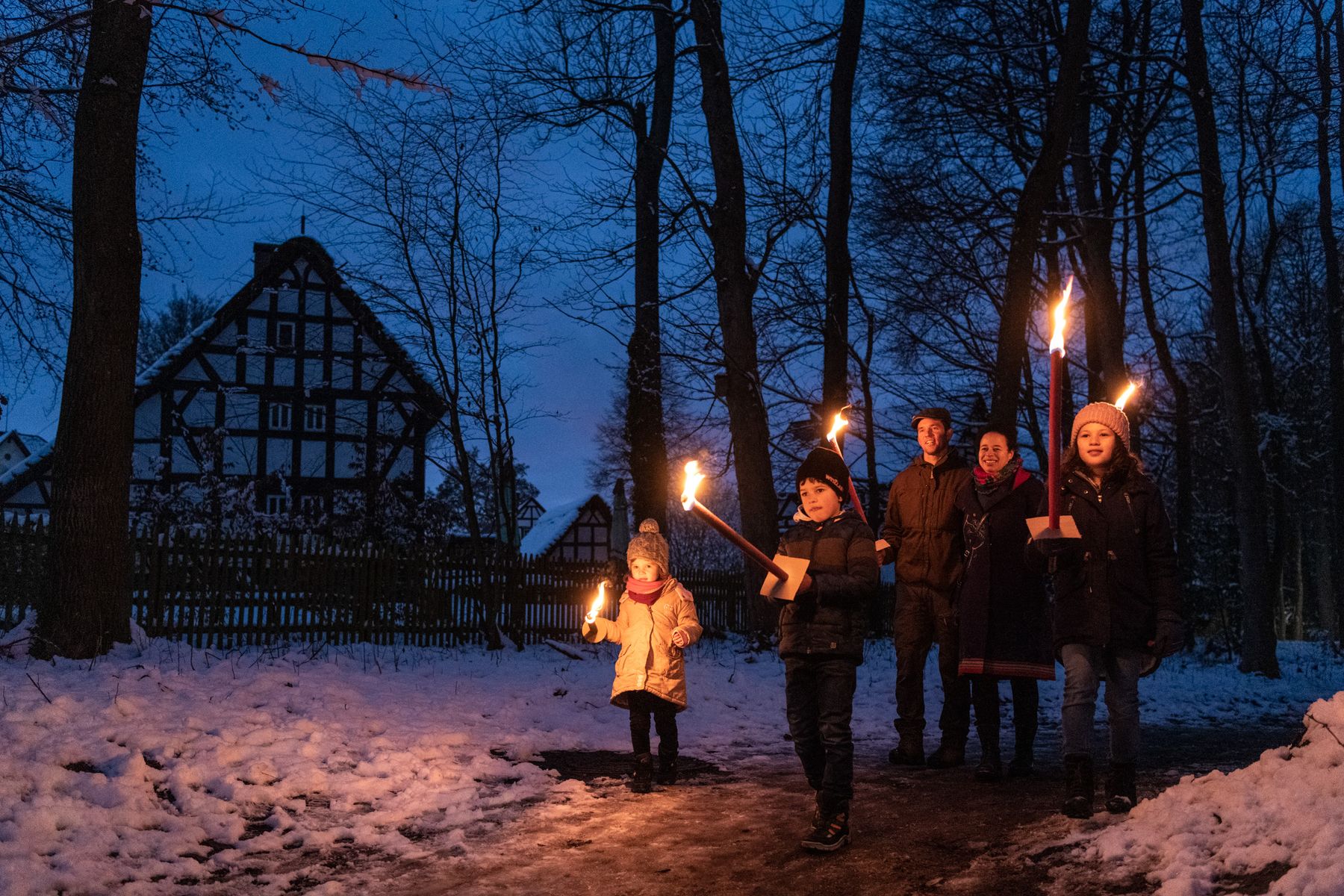 Eine Gruppe von Menschen, darunter auch Kinder, geht in der Dämmerung mit brennenden Fackeln einen verschneiten Weg entlang. Im Hintergrund sind blattlose Bäume und ein traditionelles Fachwerkhaus zu sehen.