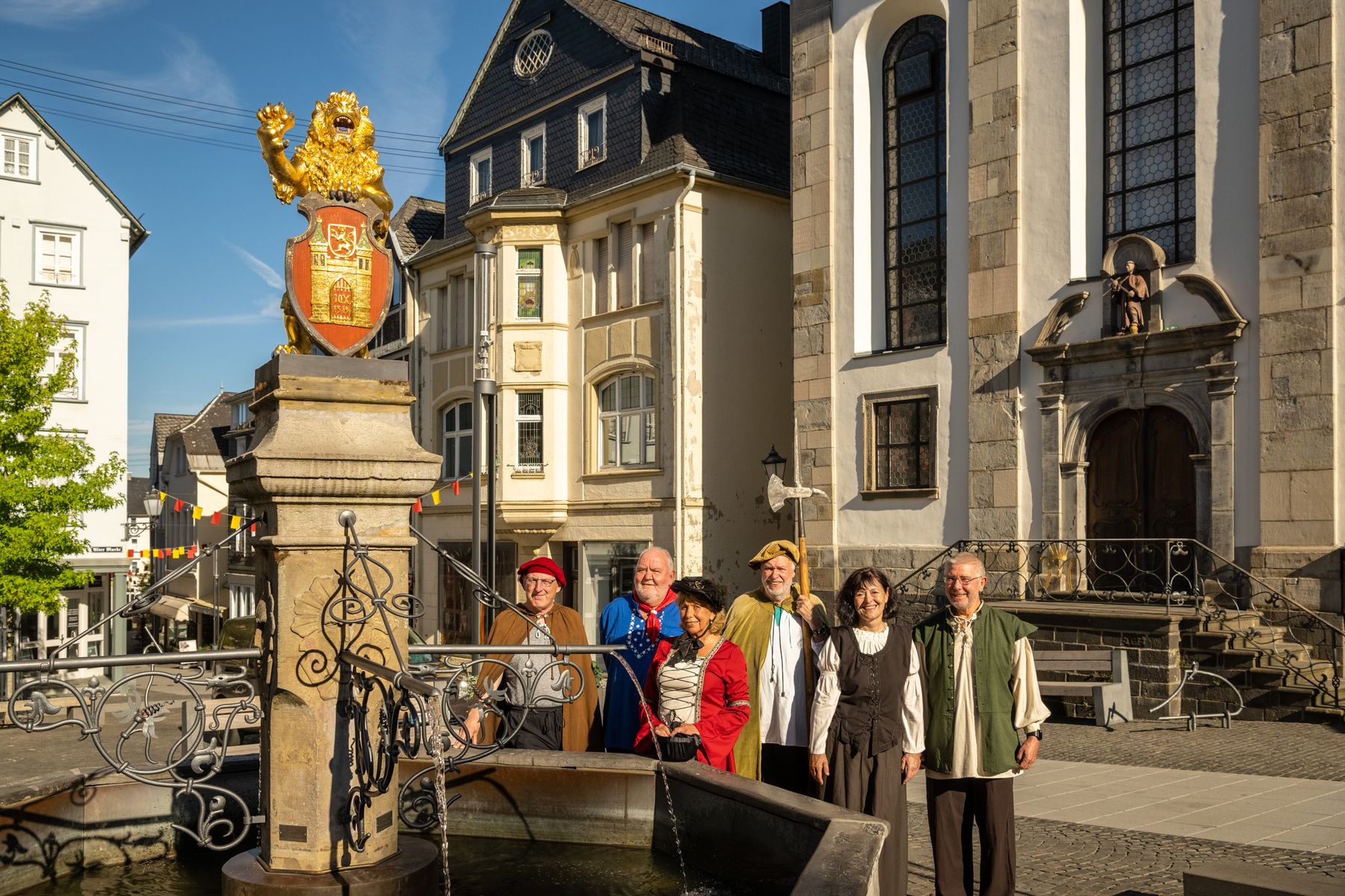 Eine Gruppe von sechs Erwachsenen in farbenfrohen, historischen Kostümen steht an einem dekorativen Brunnen auf einem europäischen Stadtplatz, mit alten Gebäuden und einer Kirche im Hintergrund an einem sonnigen Tag.