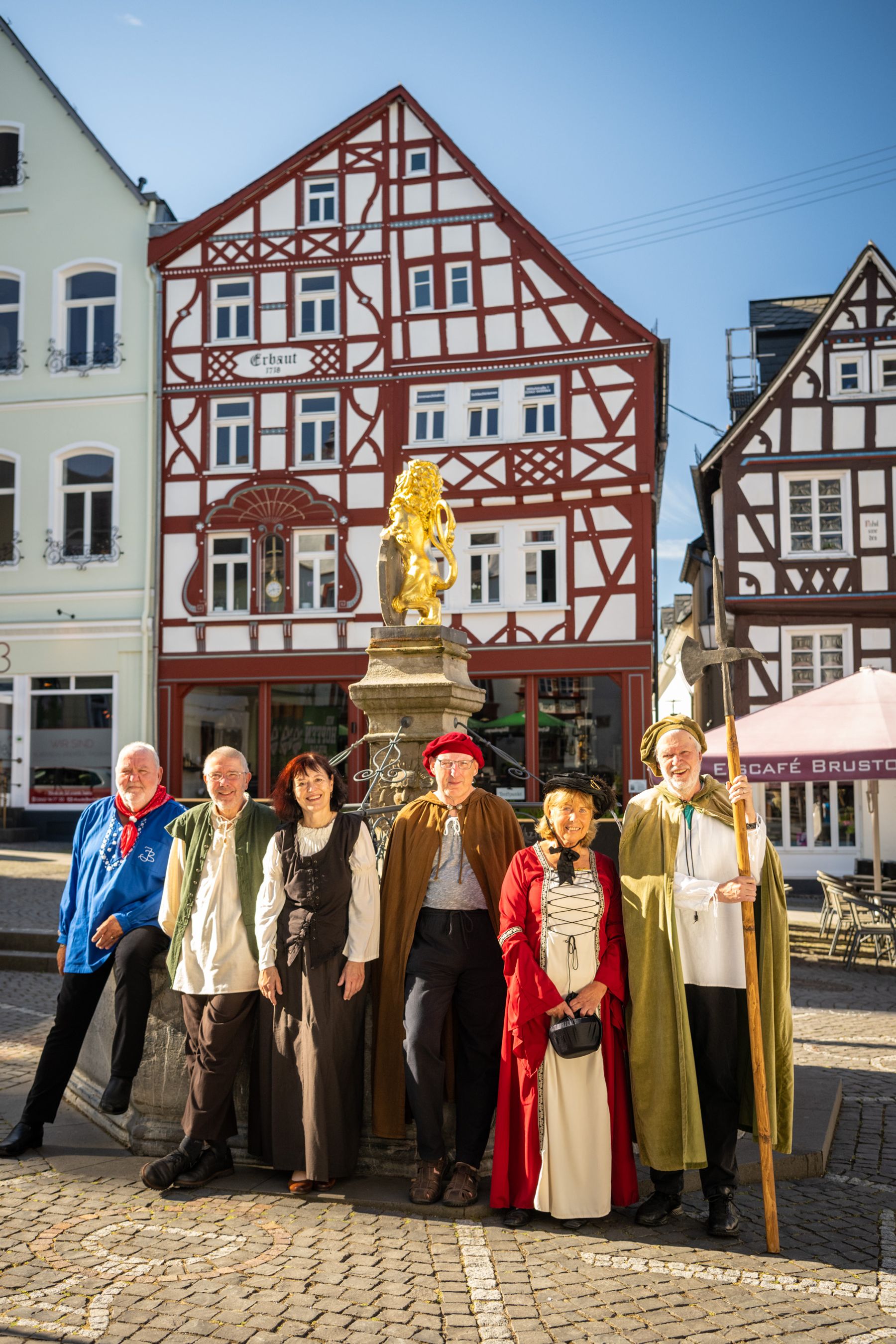 Sechs Personen in historischen Kostümen stehen lächelnd vor einem Zierbrunnen mit einer goldenen Löwenstatue, mit bunten Fachwerkhäusern im Hintergrund an einem sonnigen Tag.