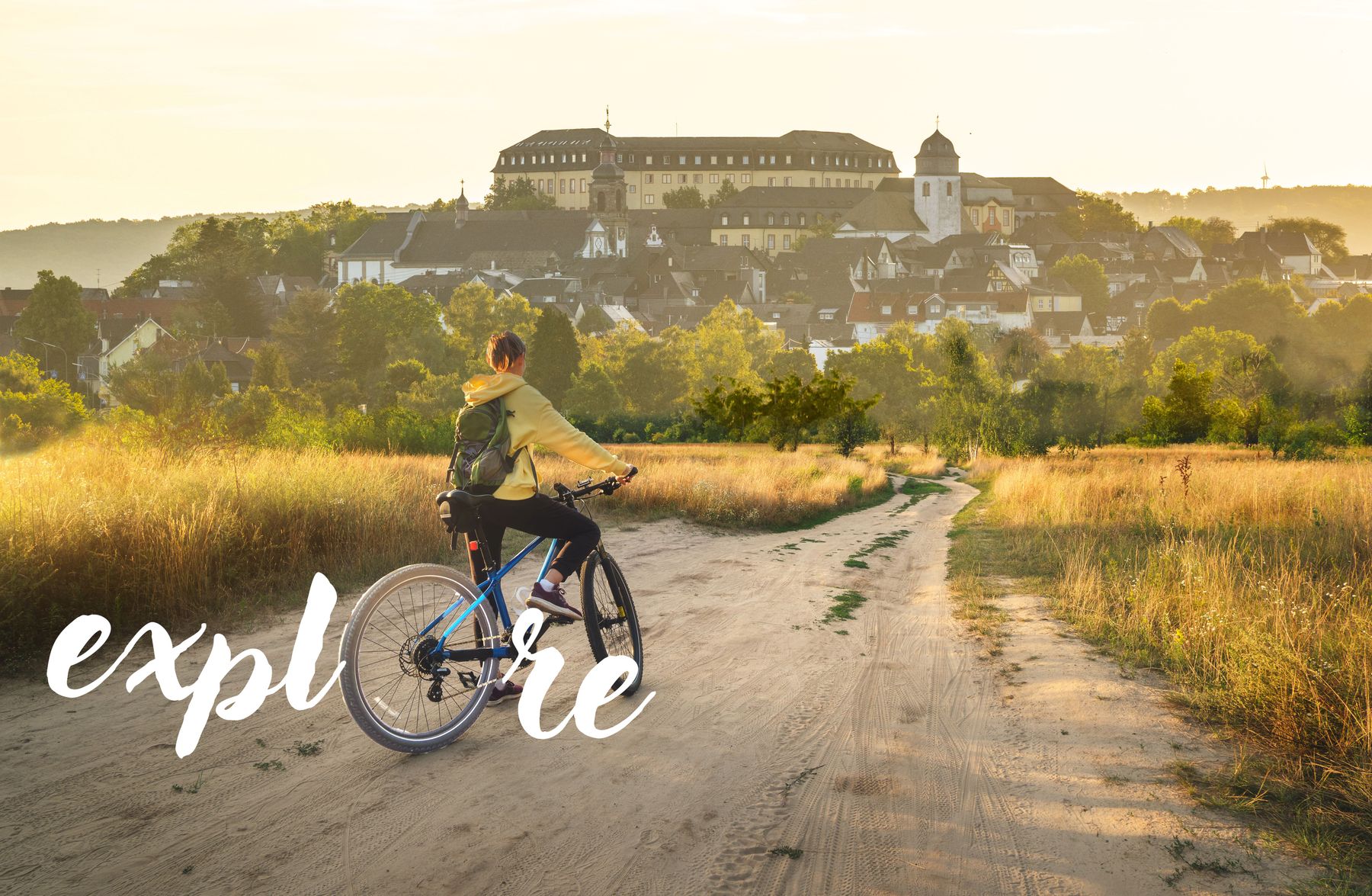 Eine Person auf einem Fahrrad fährt auf einem unbefestigten Weg durch goldene Felder auf ein malerisches Dorf und ein großes Gebäude bei Sonnenuntergang zu, wobei das Wort explore in weißer Schrift quer über das Bild geschrieben ist.
