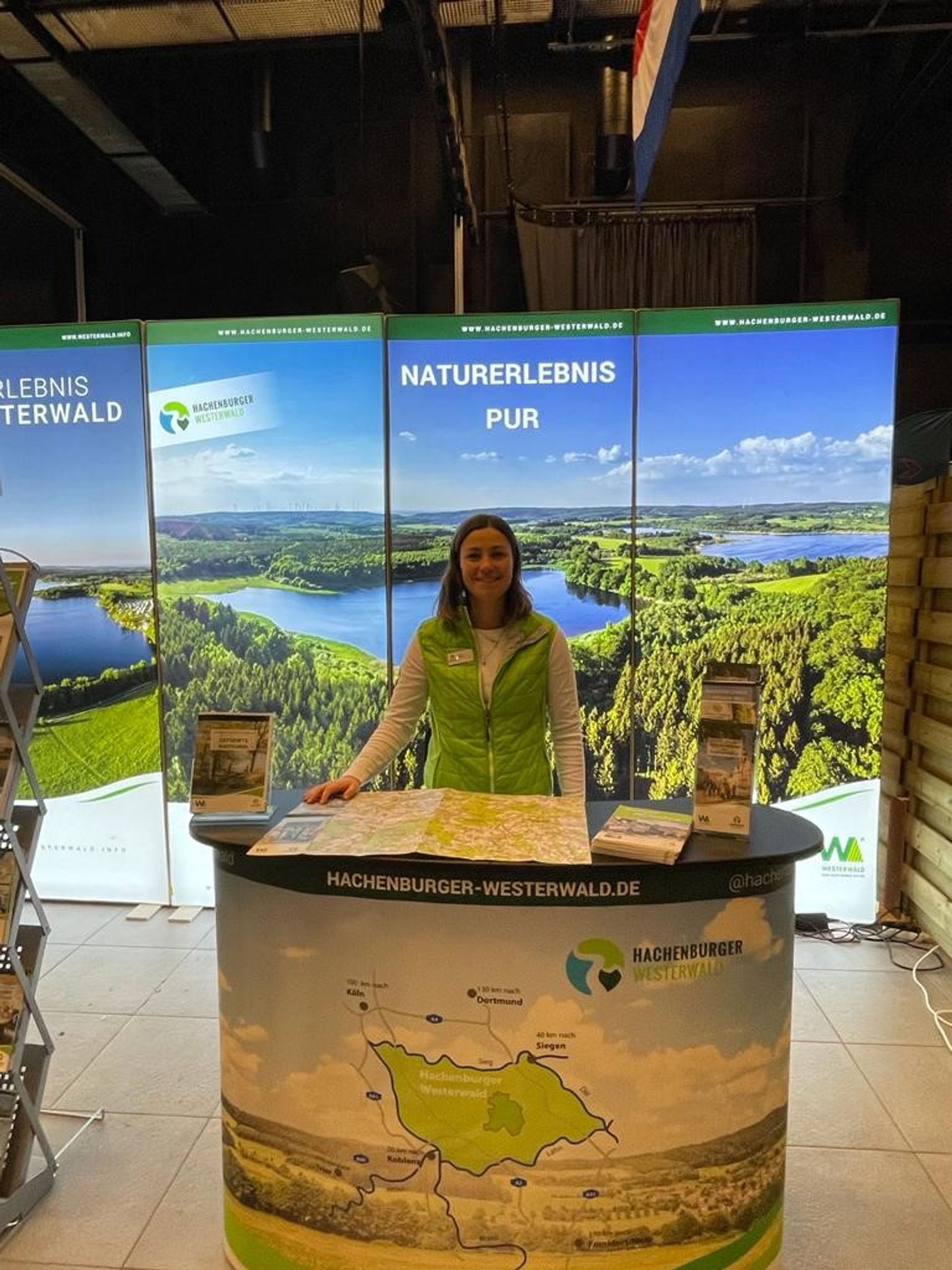 Eine Frau in grüner Weste steht hinter einem Informationsstand mit Karten und Broschüren, die für den Hachenburger Westerwald werben. Im Hintergrund sind große Landschaftstafeln und die Aufschrift NATURERLEBNIS PUR zu sehen.