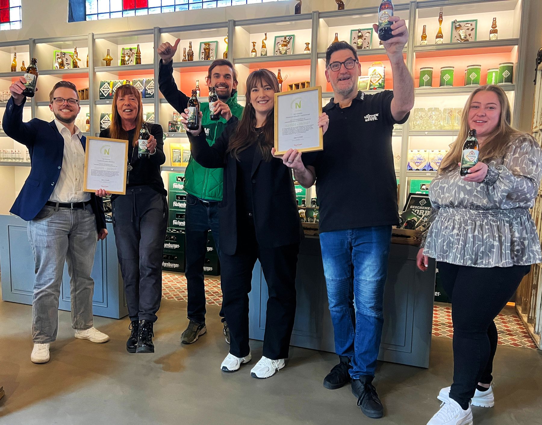 Sechs Personen stehen in einem Raum, lächeln und halten feierlich Getränke und Urkunden in die Höhe. Sie stehen vor Regalen, in denen verschiedene Flaschen und Gegenstände ausgestellt sind. Die Stimmung ist fröhlich und festlich.