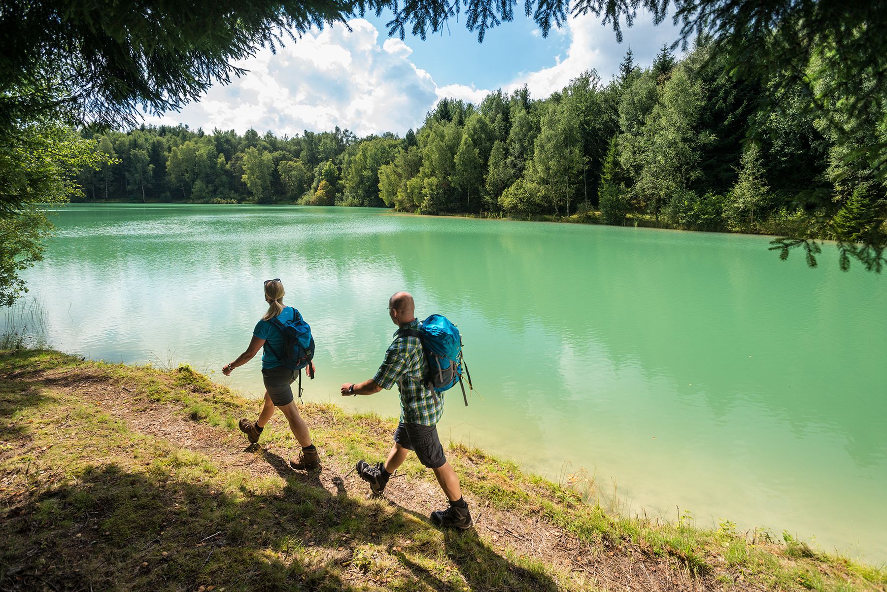 Zwei Wanderer mit Rucksäcken gehen auf einem unbefestigten Weg neben einem türkisfarbenen See, umgeben von grünen Bäumen und unter einem teilweise bewölkten Himmel.