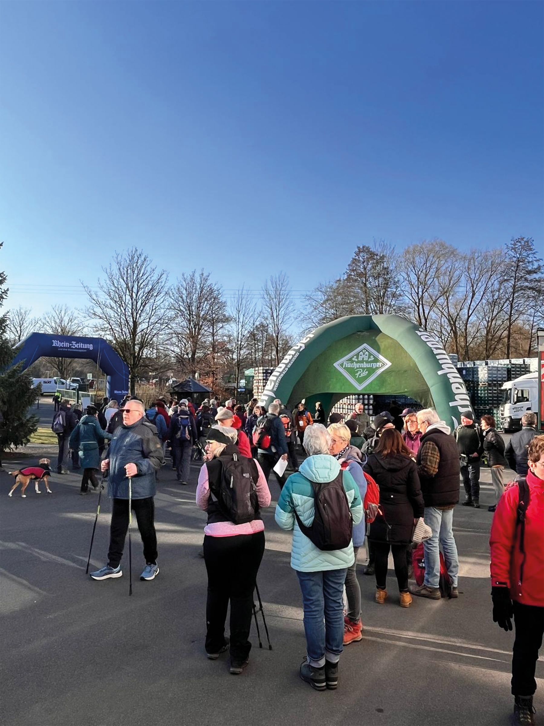 Eine Gruppe von Menschen, viele mit Rucksäcken und Wanderstöcken, versammelt sich an einem sonnigen Tag im Freien in der Nähe aufblasbarer Zelte und Veranstaltungsbanner; im Hintergrund sind einige Bäume und ein Hund zu sehen.
