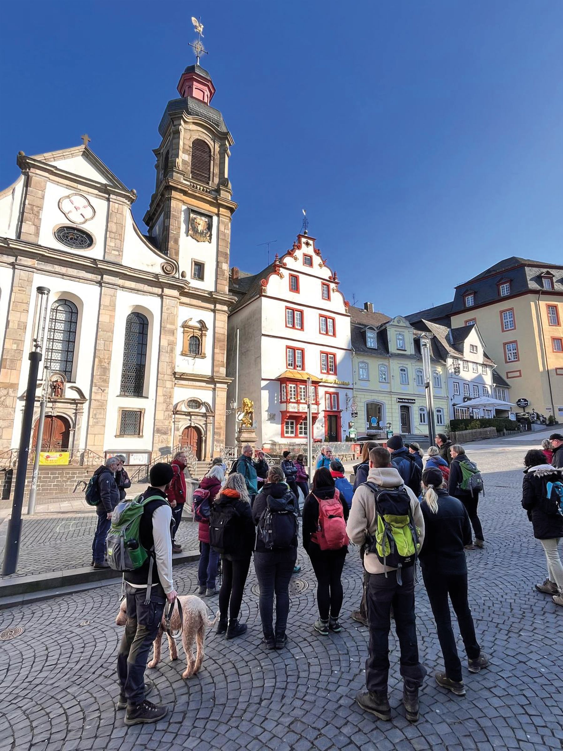 Eine Gruppe von Menschen mit Rucksäcken und einem Hund steht auf einem kopfsteingepflasterten Platz mit Blick auf historische europäische Gebäude unter einem klaren blauen Himmel. Die Szene erinnert an eine Stadtbesichtigung oder einen geführten Spaziergang.