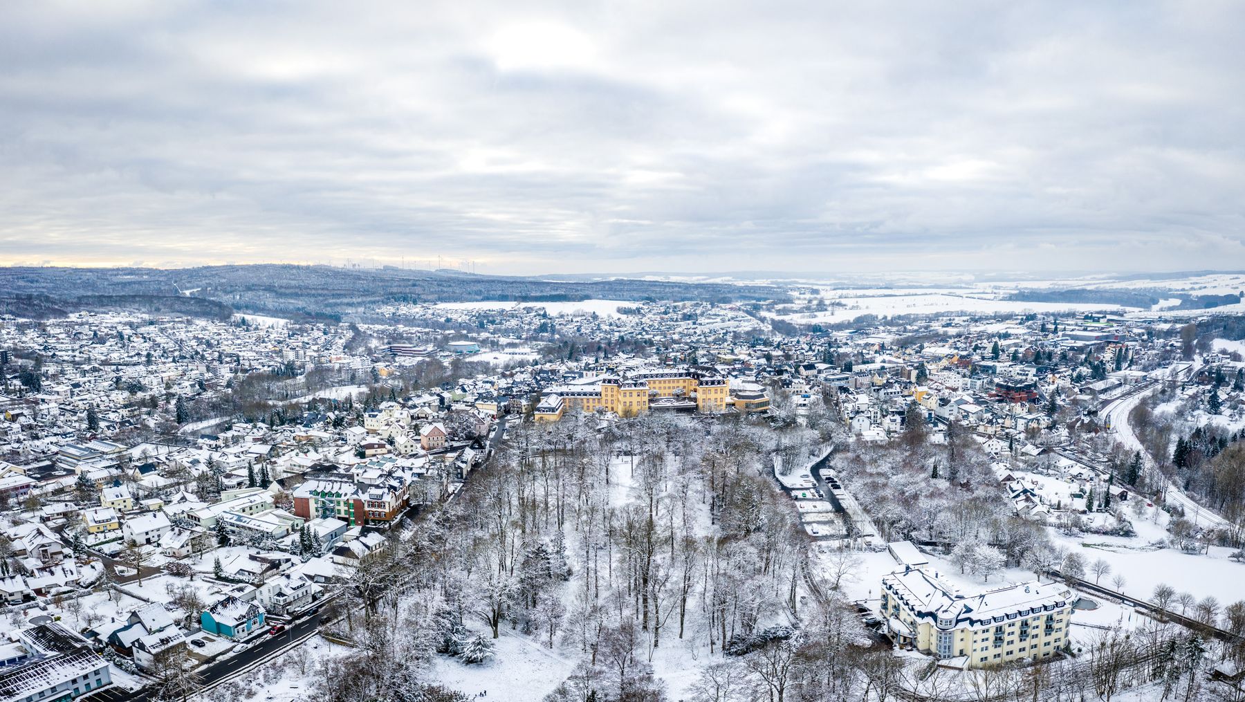 Luftaufnahme einer verschneiten Stadt mit vielen Häusern, Straßen und Bäumen. Die Landschaft ist mit Schnee bedeckt, und ein großes gelbes Gebäude ragt in der Mitte hervor. Der Himmel ist bewölkt und die Szene sieht kalt und winterlich aus.