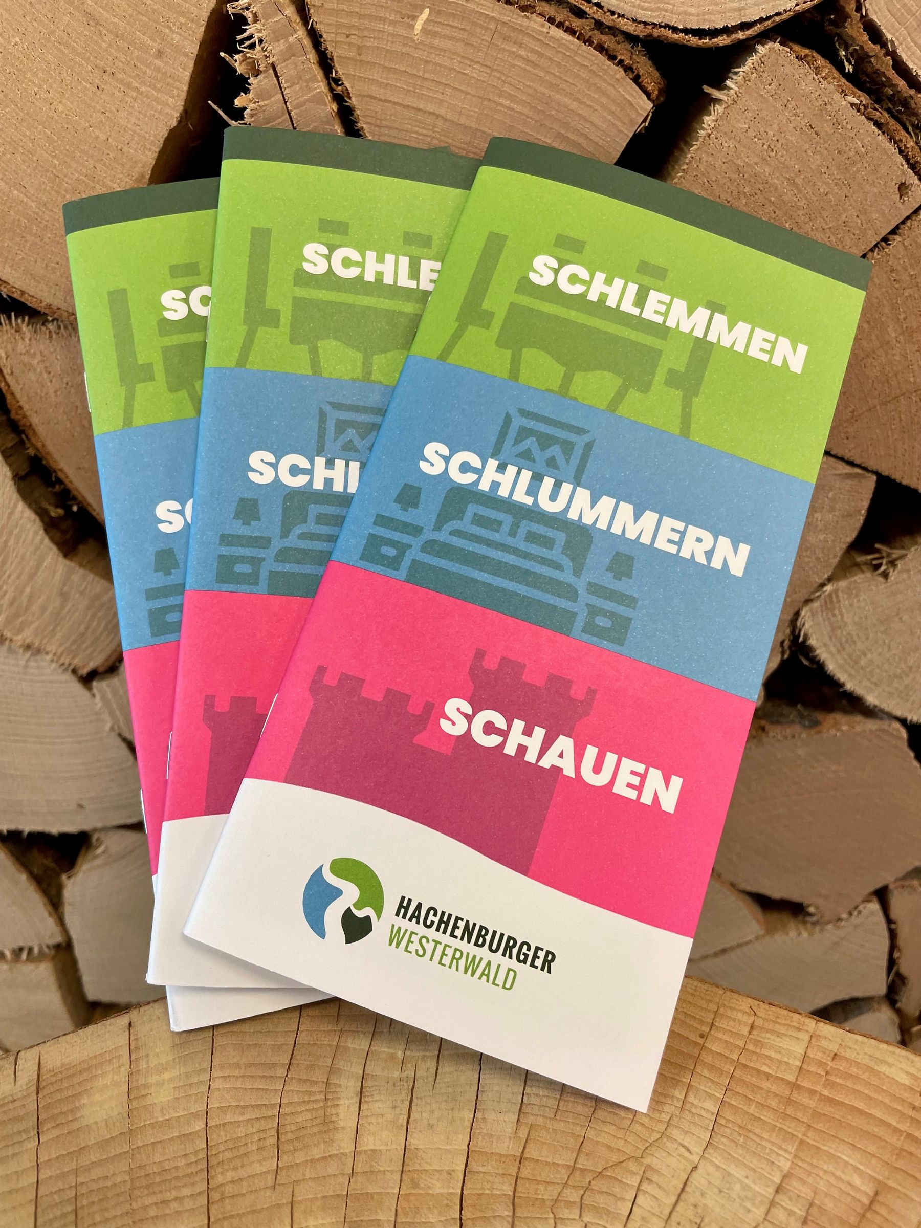 Drei bunte Flugblätter mit den Titeln Schlemmen, Schlummern und Schauen sind auf gehacktem Brennholz gestapelt. Die Flugblätter tragen am unteren Rand auch das Logo und den Namen Hachenburger Westerwald.