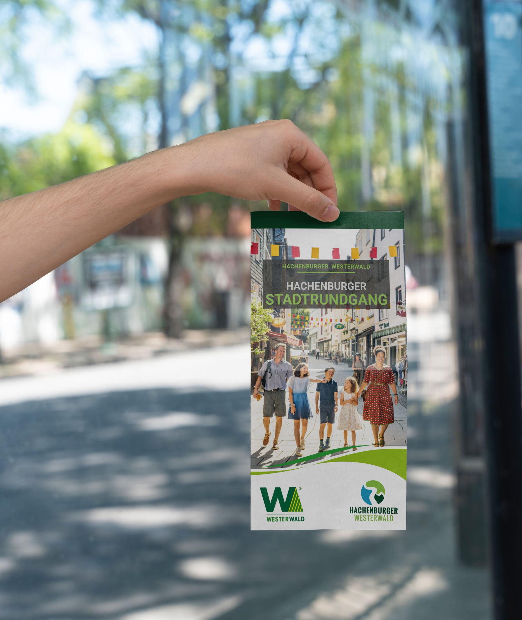 Eine Hand hält eine farbenfrohe Broschüre für den Hachenburger Stadtrundgang, die eine Familie zeigt, die in einer lebendigen Stadtkulisse spazieren geht, wobei Bäume und Gebäude im Hintergrund verschwimmen.