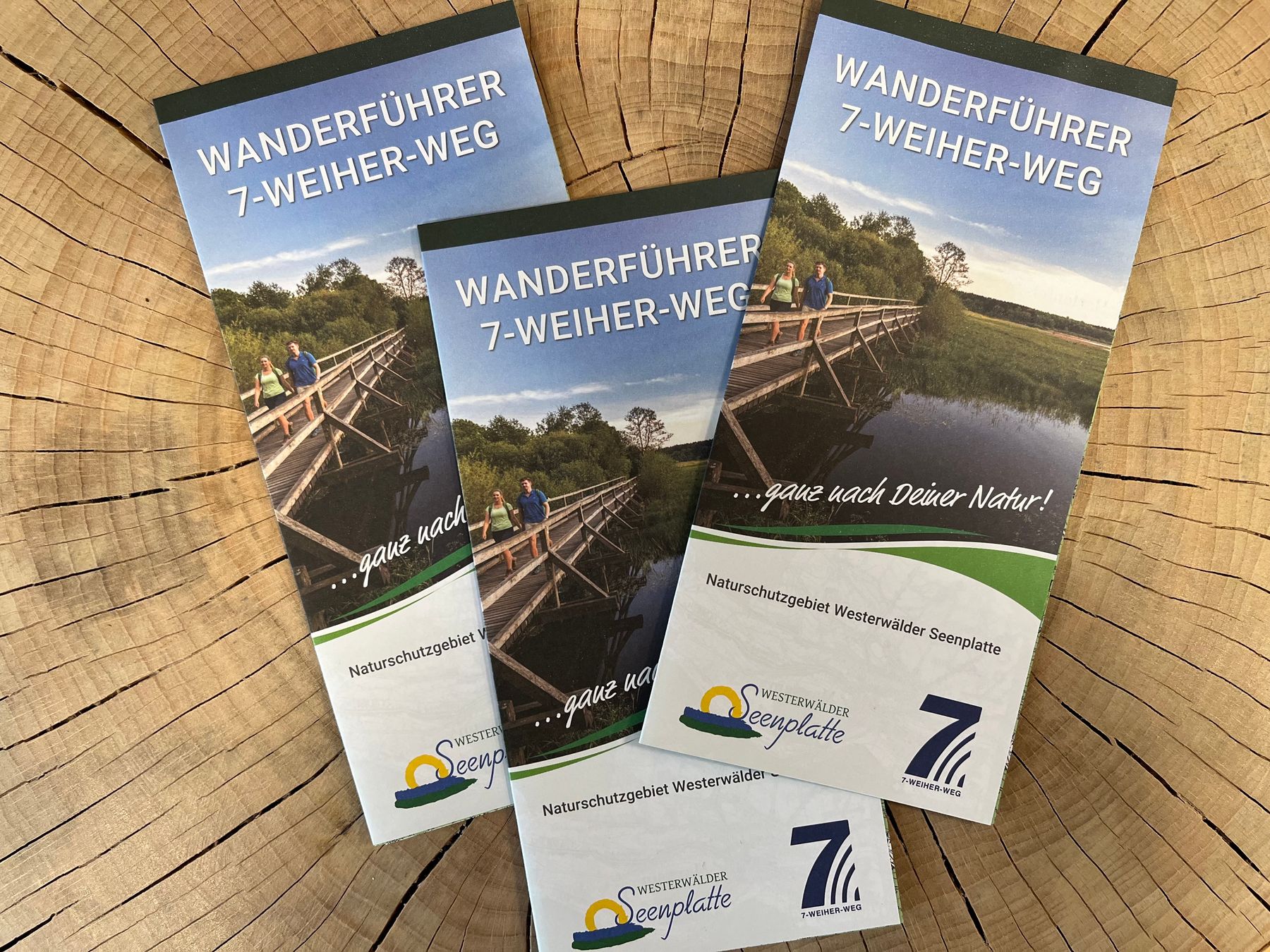 Drei Broschüren mit dem Titel WANDERFÜHRER 7-WEIHER-WEG sind auf der Oberfläche eines Baumstamms angebracht und zeigen ein Foto von zwei Personen auf einer Holzbrücke über das Wasser, mit der Natur im Hintergrund.