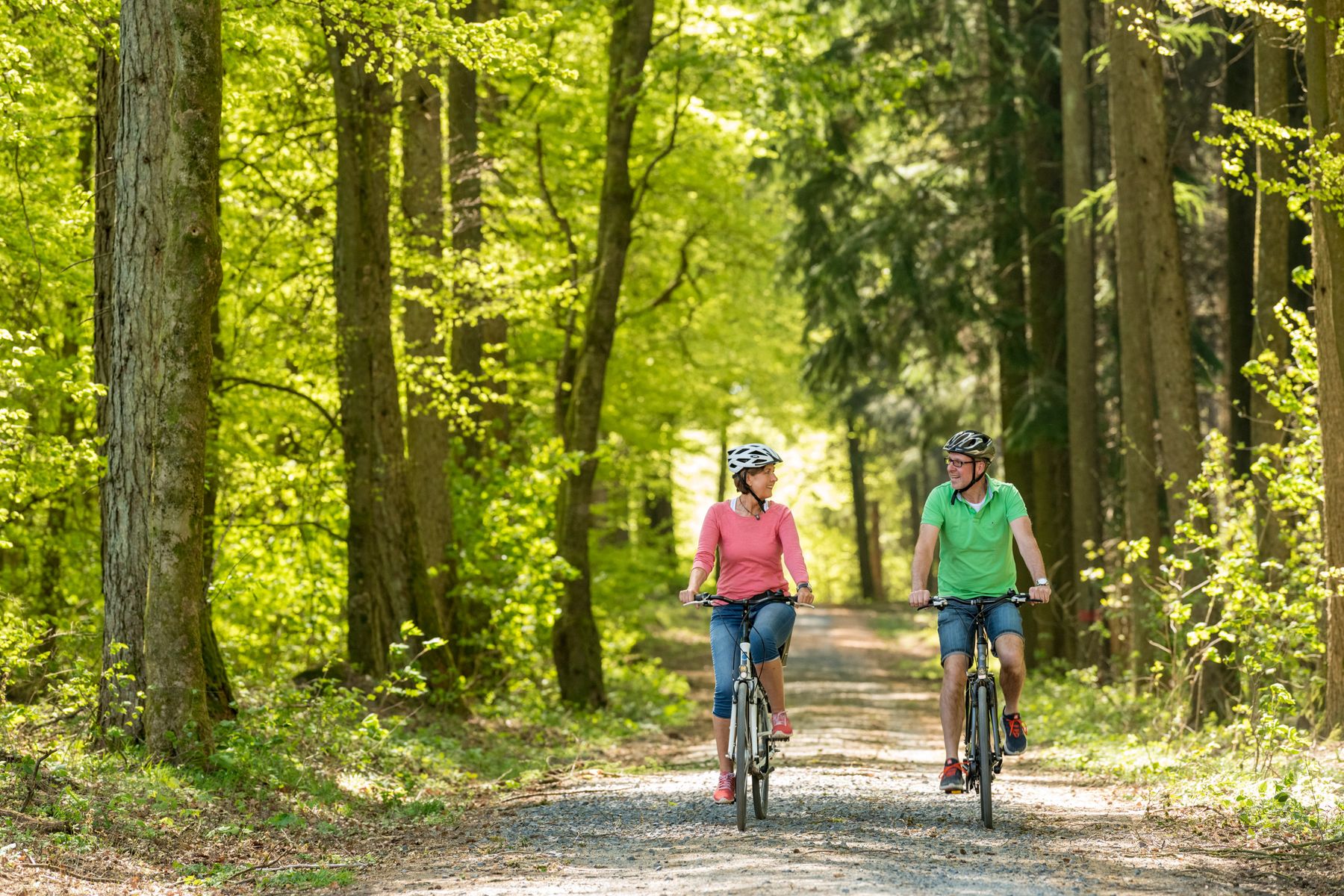 Ein Mann und eine Frau fahren mit dem Fahrrad auf einem unbefestigten Weg durch einen sonnenbeschienenen, grünen Wald. Beide tragen Helme und Freizeitkleidung und genießen die Natur und die Gesellschaft des anderen, während sie Seite an Seite radeln.