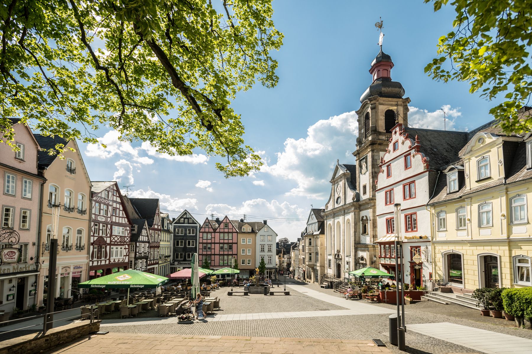 Ein malerischer europäischer Stadtplatz mit farbenfrohen, historischen Gebäuden, Cafés mit Sitzgelegenheiten im Freien unter grünen Sonnenschirmen, Kopfsteinpflasterstraßen und einer hohen Kirche mit einem Uhrenturm unter einem hellen, teilweise bewölkten Himmel.