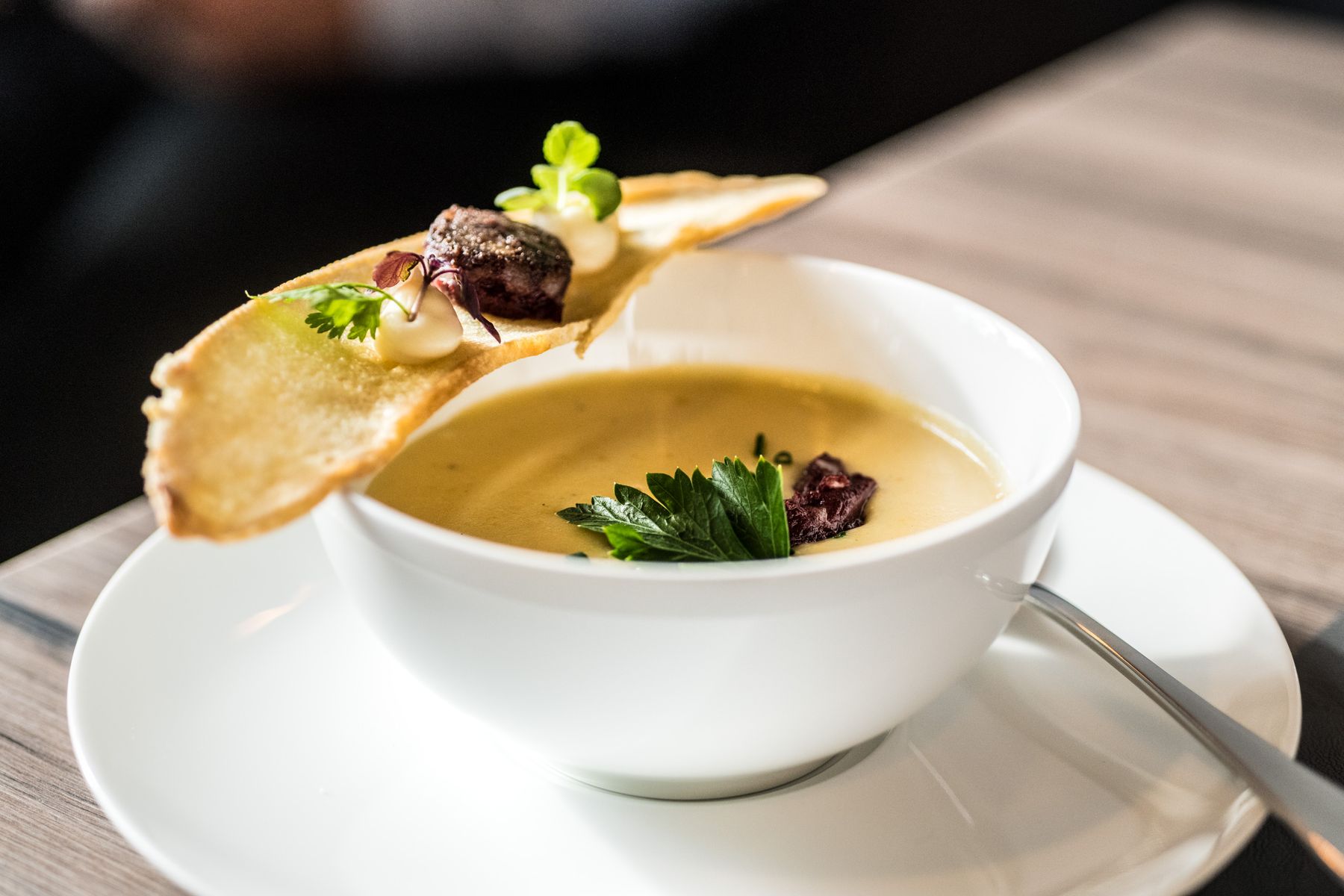 Eine weiße Schale mit cremiger Suppe, garniert mit Petersilie, serviert mit einer getoasteten Brotscheibe, die mit Kräutern und Saucenklecksen belegt ist, auf einem Holztisch, wobei der Löffel auf dem Rand der Schale ruht.