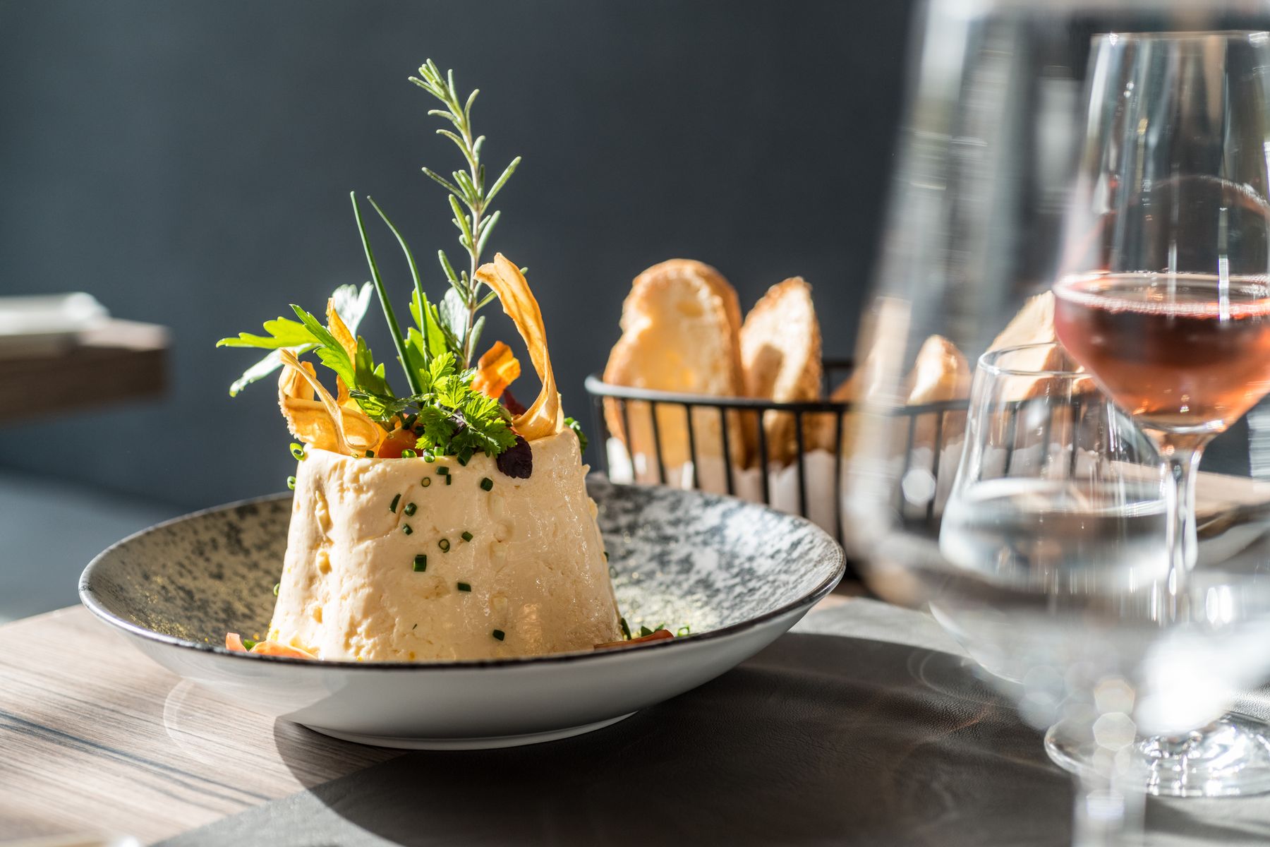 Eine plattierte Gourmet-Vorspeise mit frischen Kräutern und Garnierungen, serviert auf einer modernen grauen Schale. Im Hintergrund sind ein Korb mit Brotscheiben und ein Glas Roséwein auf einem sonnenbeschienenen Tisch zu sehen.