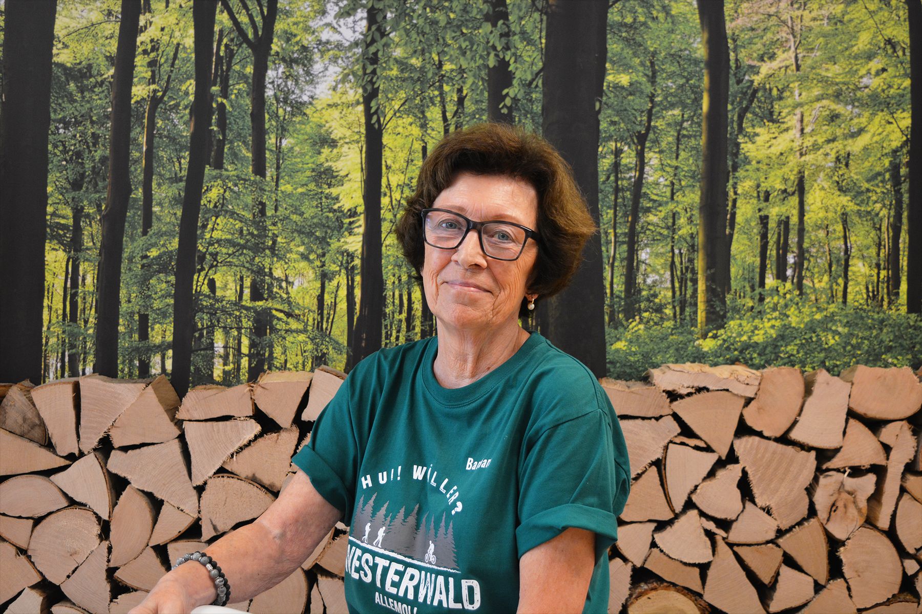 Eine Frau mit kurzen braunen Haaren und Brille, die ein grünes Westerwald-T-Shirt trägt, sitzt vor einem Stapel Brennholz mit einem Waldhintergrund.