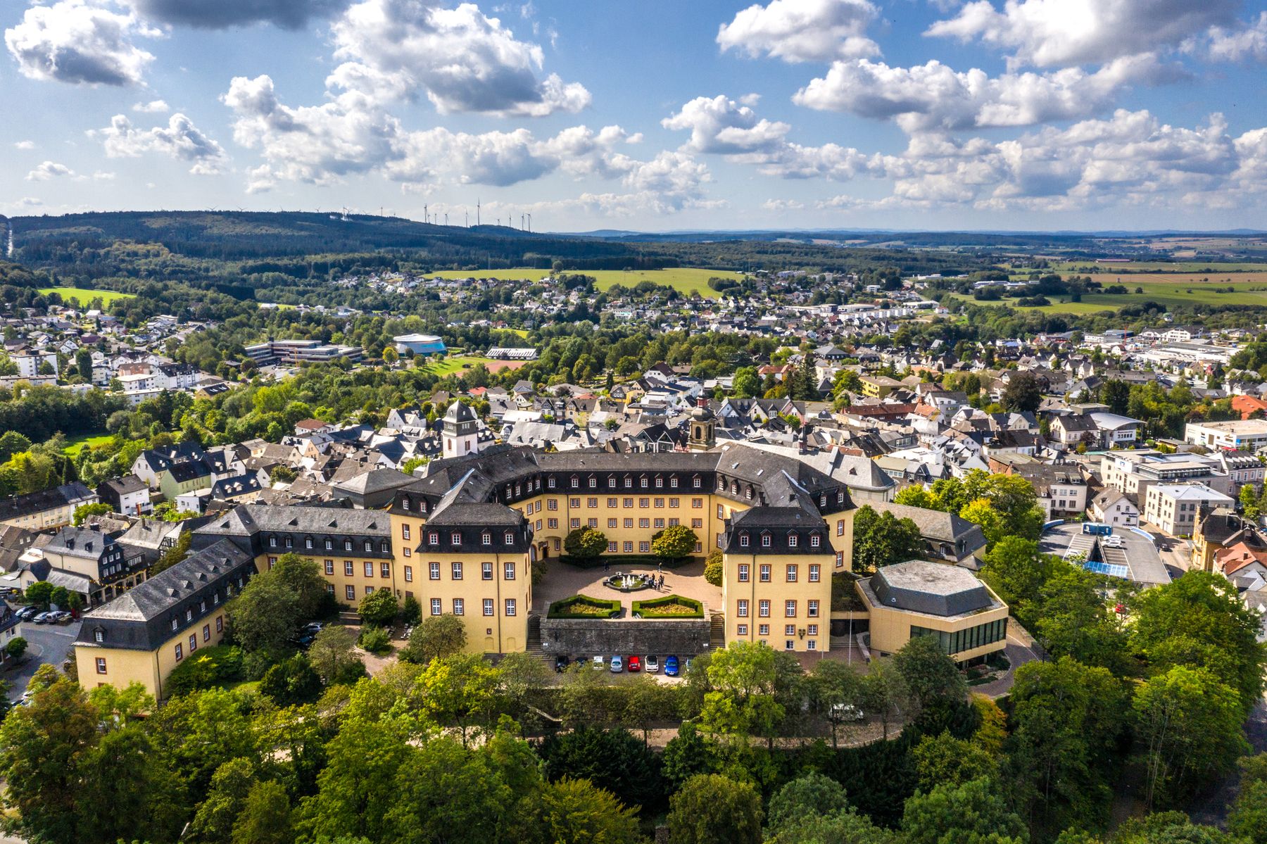 Luftaufnahme eines großen, historischen, gelben Gebäudes mit Innenhof und umliegenden Bäumen in einer kleinen Stadt mit Häusergruppen und sanften Hügeln unter einem bewölkten Himmel.