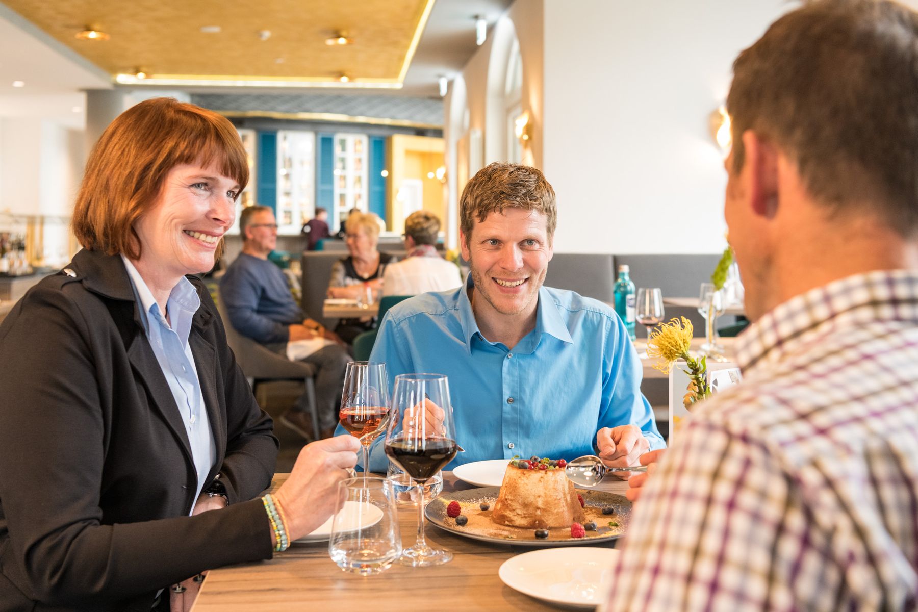 Drei Personen sitzen an einem Restauranttisch, lächeln und genießen gemeinsam das Dessert. Zwei halten Weingläser, und auf dem Tisch steht ein Kuchen. Im Hintergrund sind in einer gut beleuchteten, modernen Umgebung weitere Gäste zu sehen.