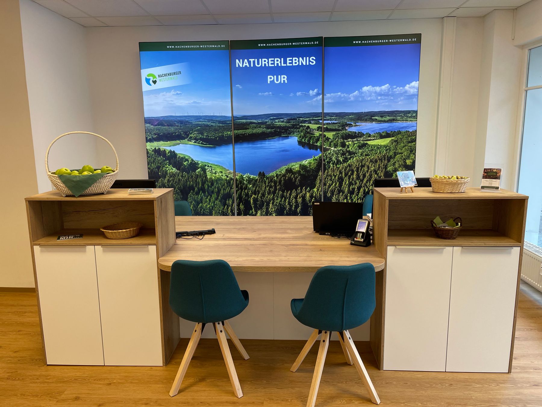 Ein moderner Empfangsbereich mit zwei blaugrünen Stühlen an einem hölzernen Schreibtisch, Körben mit Äpfeln auf der Theke und einem großen Wanddisplay, das einen malerischen Wald und See mit dem Text NATURERLEBNIS PUR zeigt.