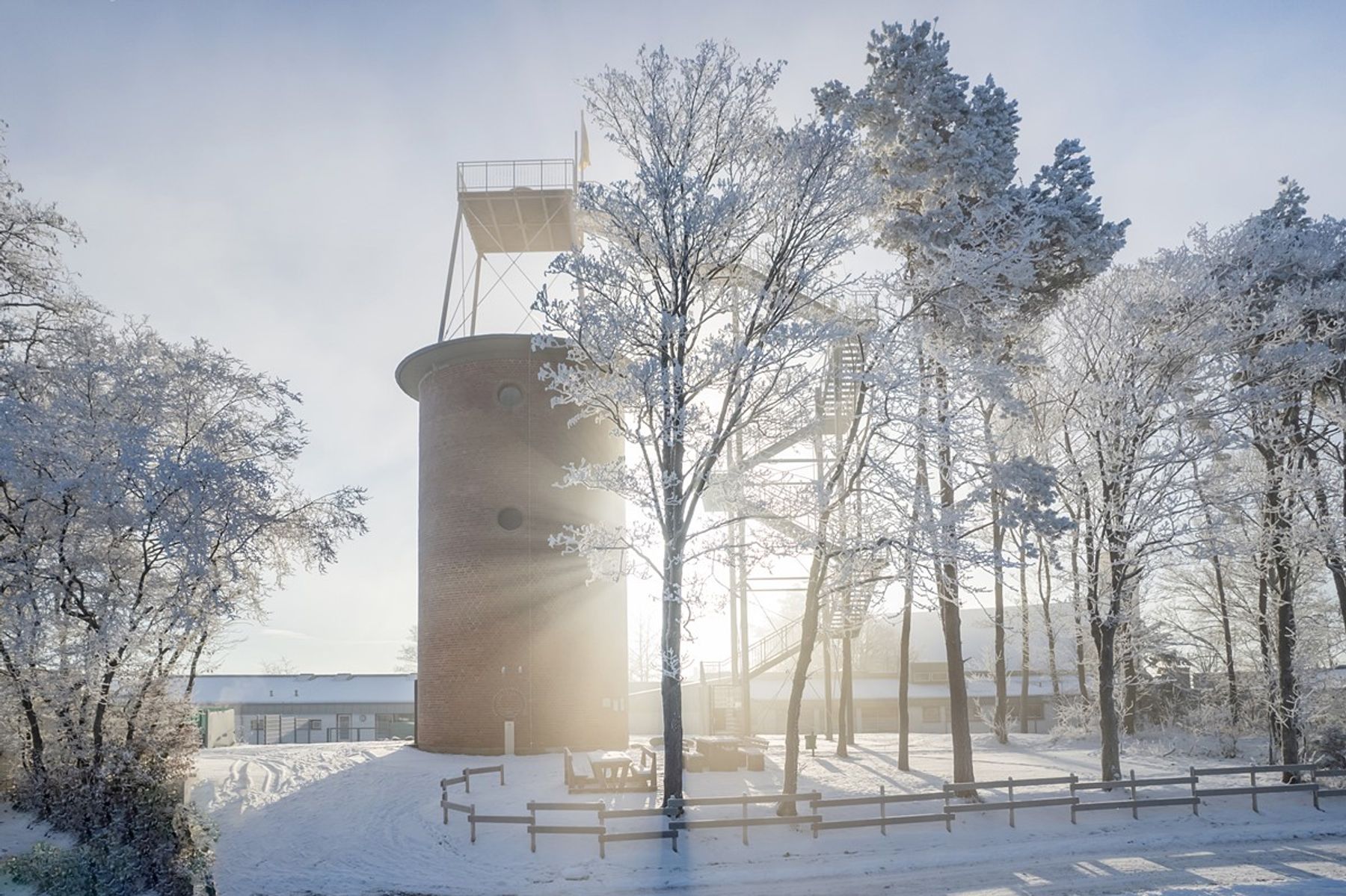 Ein runder Backsteinturm steht an einem hellen Wintertag zwischen schneebedeckten Bäumen. Das Sonnenlicht fällt durch die Äste und wirft lange Schatten auf den Schnee. In der Nähe sind ein Holzzaun und ein Gebäude zu sehen.