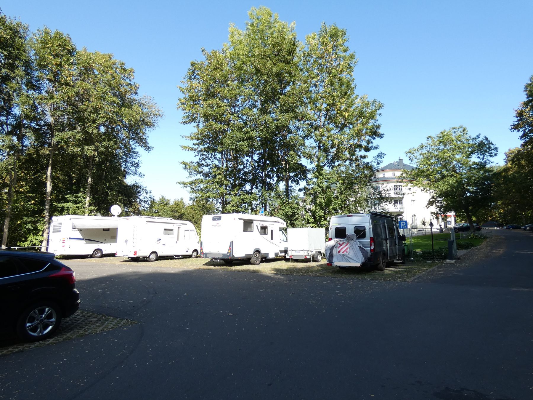 Mehrere Wohnmobile und ein Wohnwagen sind auf einem asphaltierten Grundstück geparkt, das von hohen grünen Bäumen umgeben ist. Im Hintergrund ist ein weißes Gebäude unter einem klaren blauen Himmel zu sehen. Ein schwarzes Auto ist teilweise auf der linken Seite zu sehen.