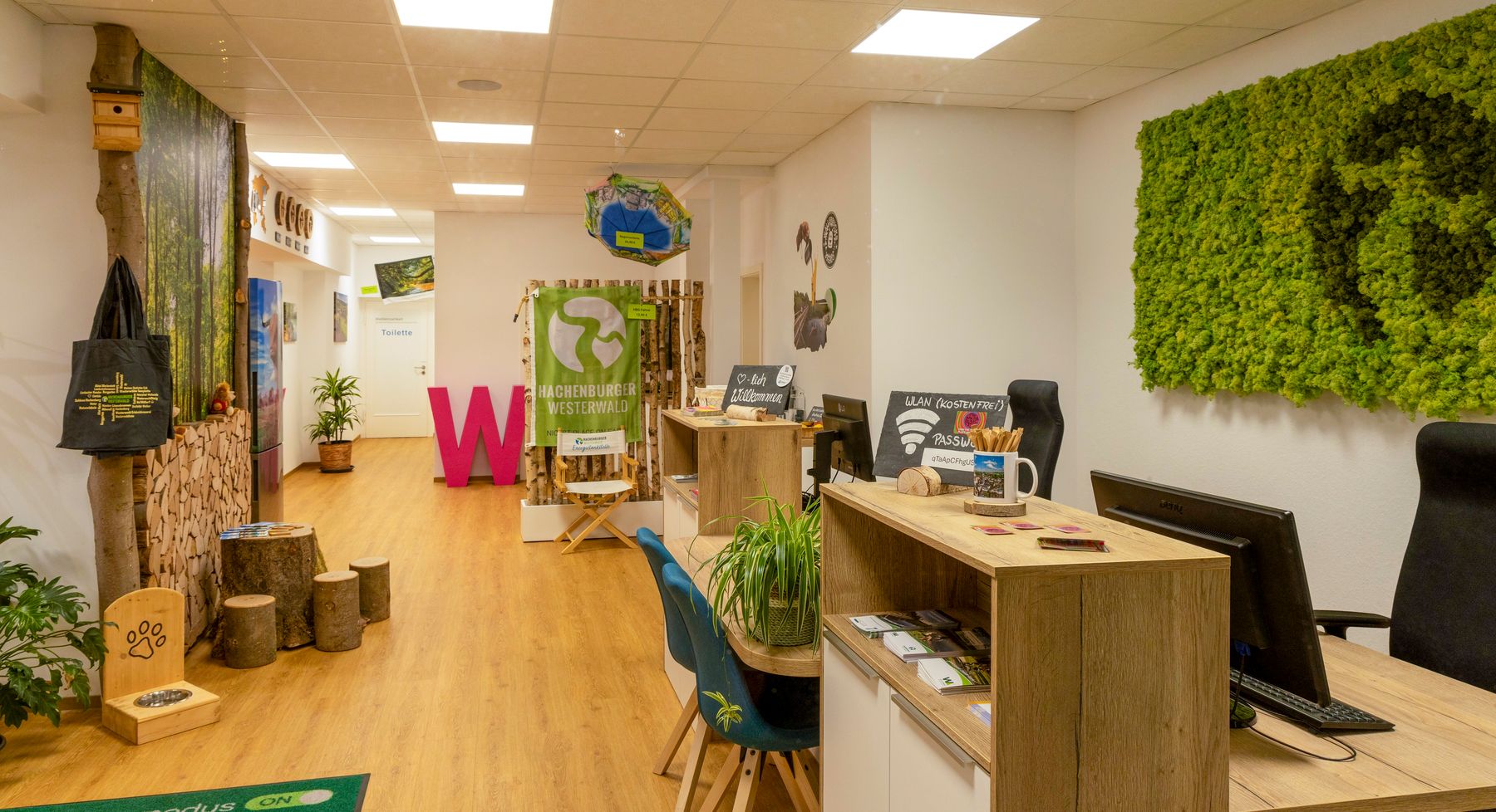 Ein modernes Büro mit Holzböden, grünem Moos an der Wand, Pflanzen, Holzakzenten, Schreibtischen mit Computern und umweltfreundlicher Dekoration, das einen von der Natur inspirierten, nachhaltigen Arbeitsbereich schafft.