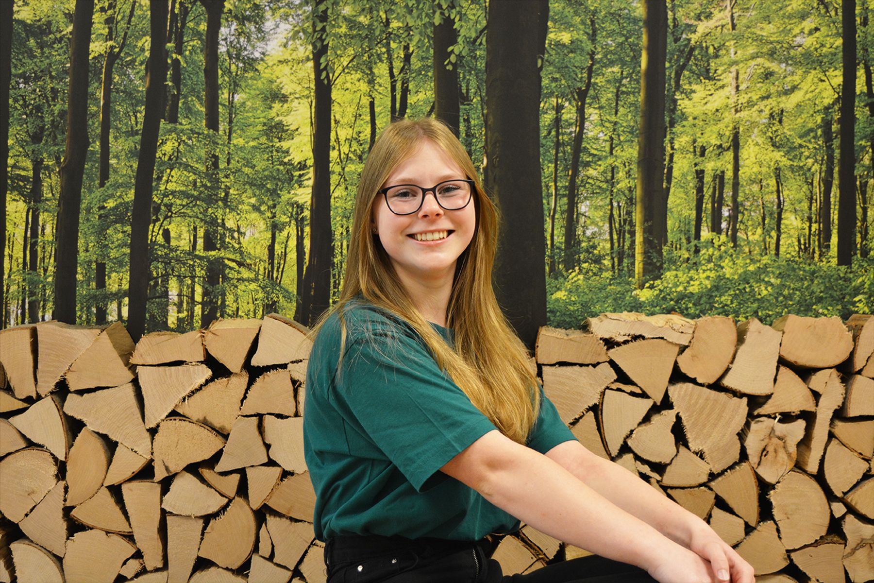 Eine junge Frau mit langen blonden Haaren und Brille sitzt lächelnd vor gestapeltem Brennholz vor einem grünen Waldhintergrund. Sie trägt ein grünes Hemd und eine schwarze Hose.