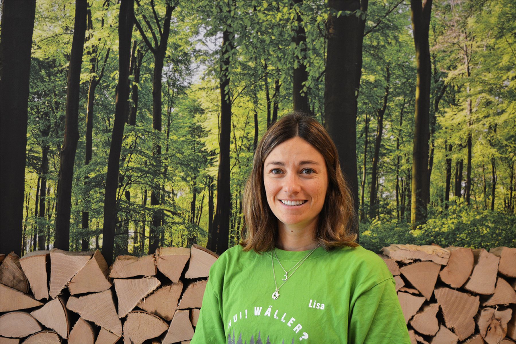 Eine Frau mit braunem Haar, die ein grünes Hemd mit der Aufschrift Oui! Wäller? und eine Halskette trägt, steht lächelnd vor einer Kulisse aus grünen Bäumen und gestapeltem Brennholz.