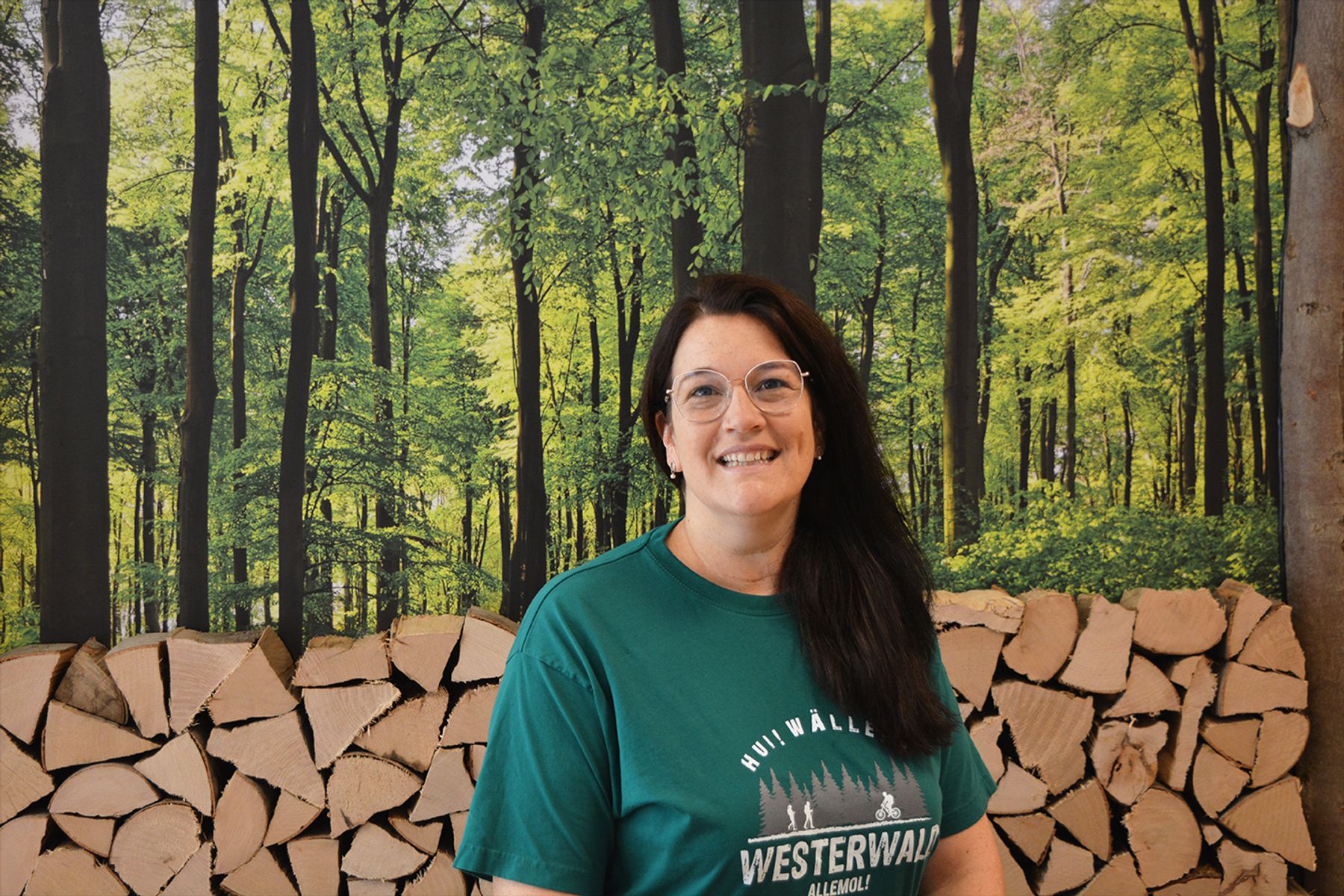 Eine Frau mit langen dunklen Haaren und Brille steht lächelnd vor gestapeltem Brennholz und einer waldähnlichen Kulisse mit hohen grünen Bäumen. Sie trägt ein grünes Westerwald-T-Shirt.