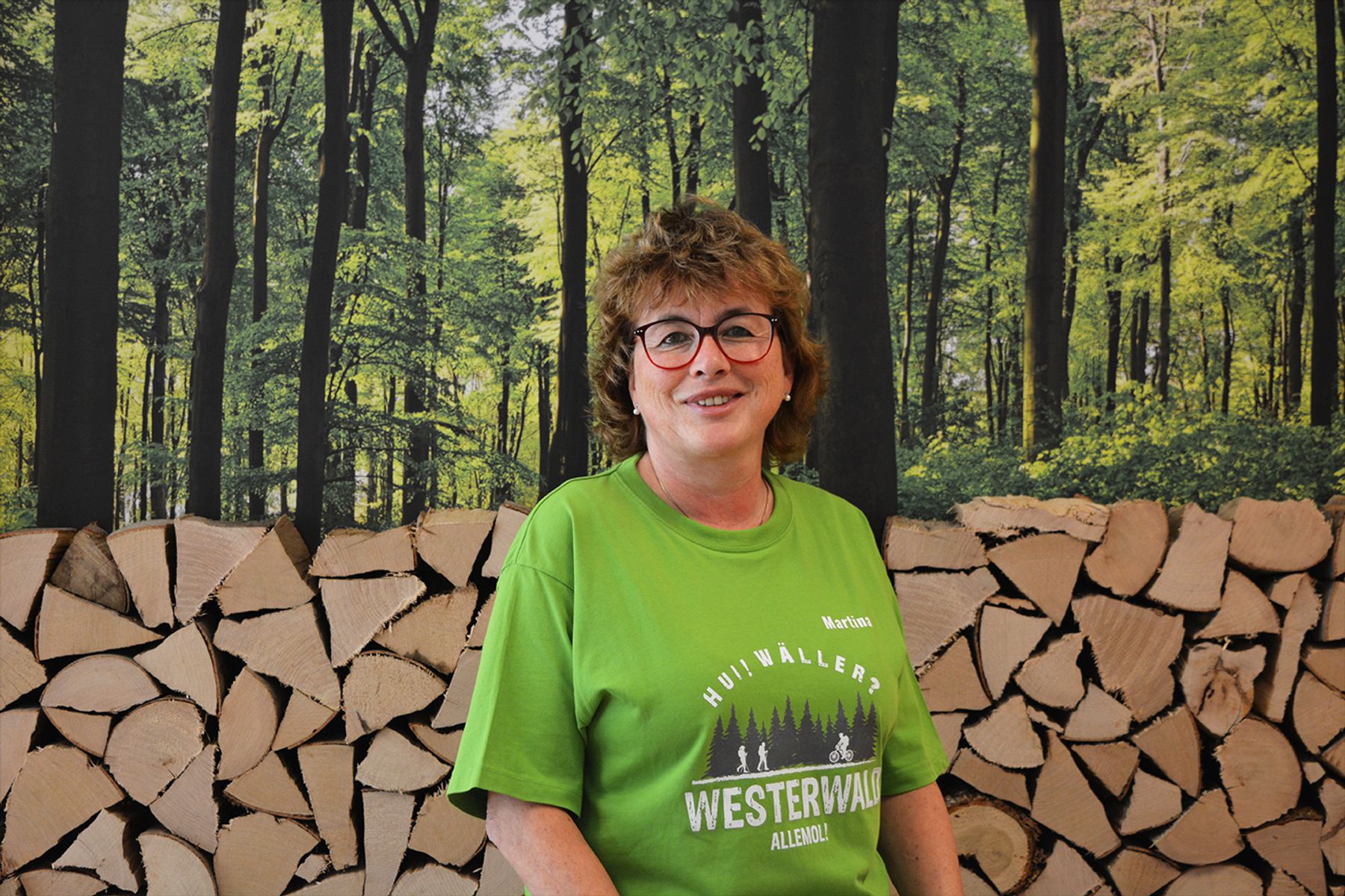 Eine Frau mit kurzen braunen Haaren und Brille, die ein leuchtend grünes T-Shirt trägt, steht vor einem Stapel Brennholz mit einem Waldwandbild im Hintergrund.