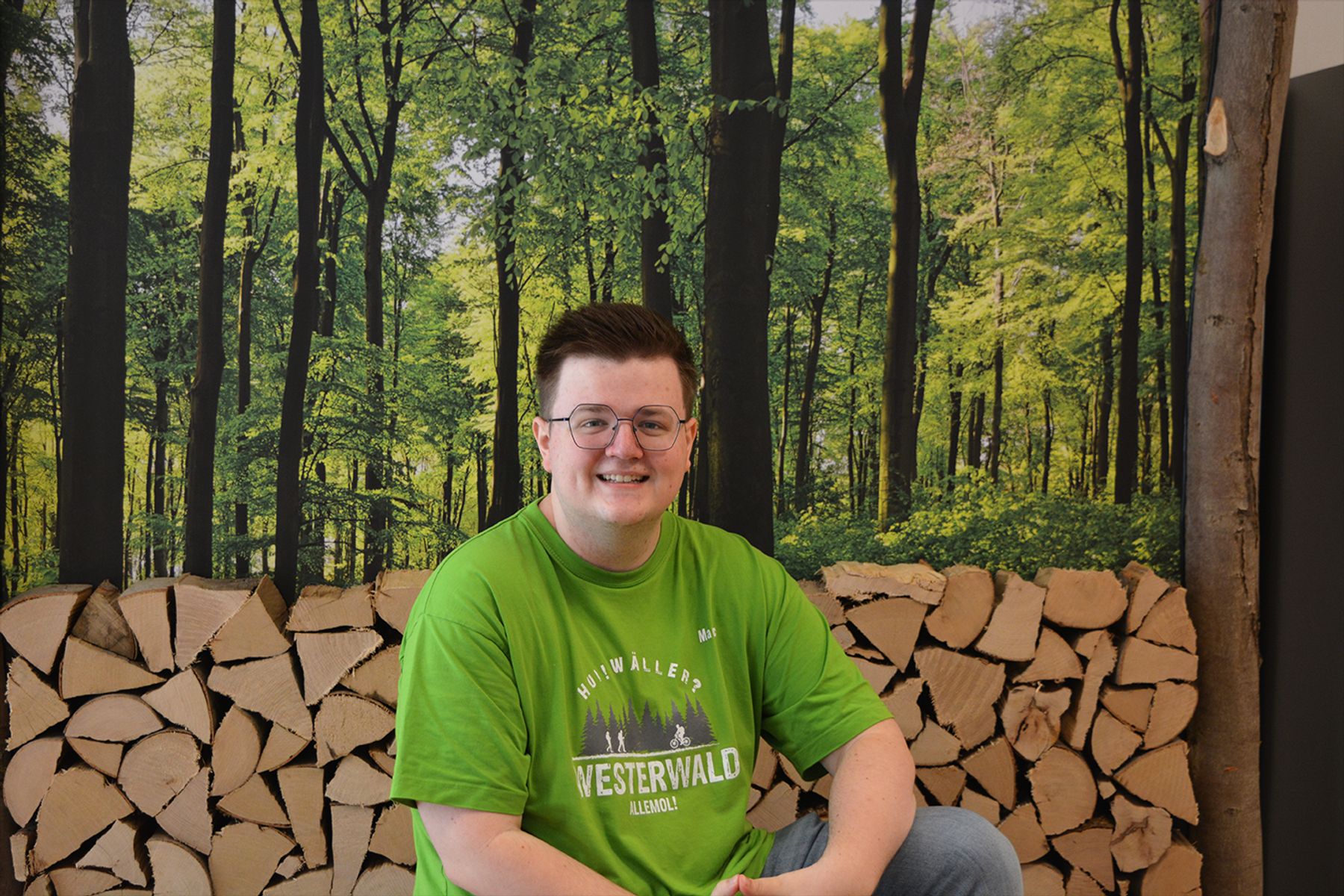 Eine lächelnde Person mit Brille und leuchtend grünem Westerwald-T-Shirt sitzt vor einem Stapel ordentlich aufgeschichtetem Brennholz, im Hintergrund ein üppig grüner Wald.