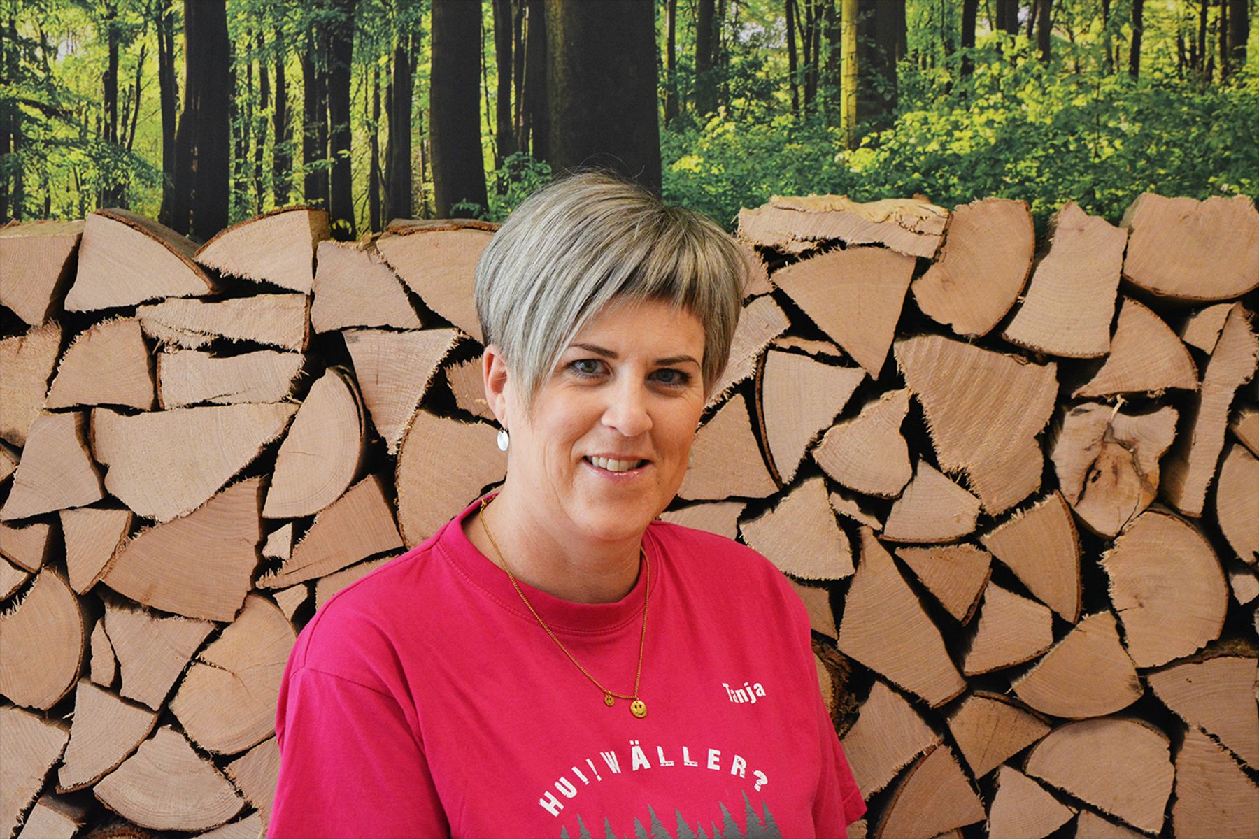 Eine Frau mit kurzen grauen Haaren, rosafarbenem Hemd und goldenen Ketten steht vor einem ordentlich aufgestapelten Stapel gehackten Brennholzes vor einem Waldhintergrund.