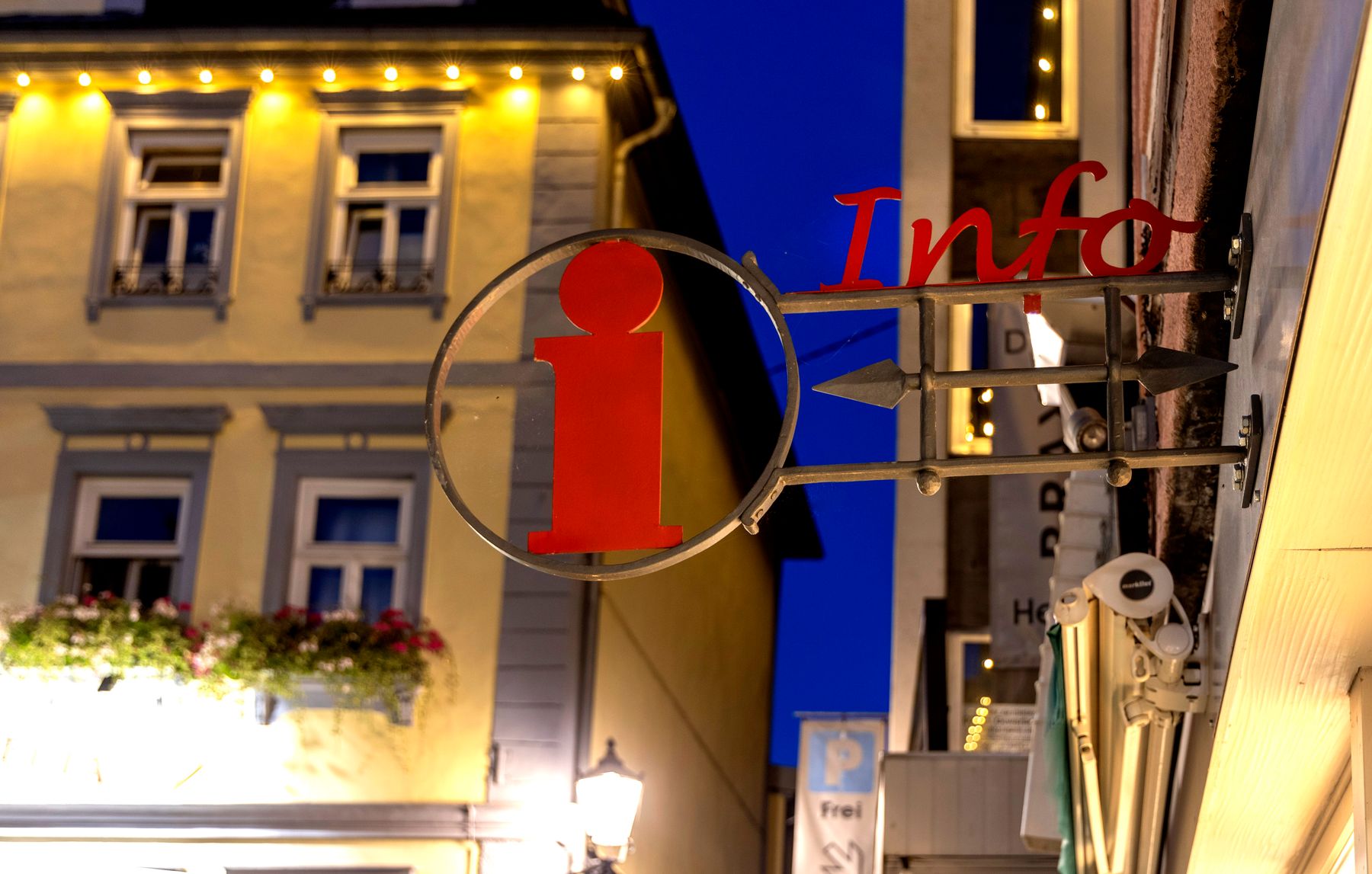 Ein rotes Info-Schild mit einem kleinen i-Symbol hängt an einer Metallhalterung vor einem Gebäude in der Abenddämmerung, mit beleuchteten Fenstern und Blumenkästen im Hintergrund.