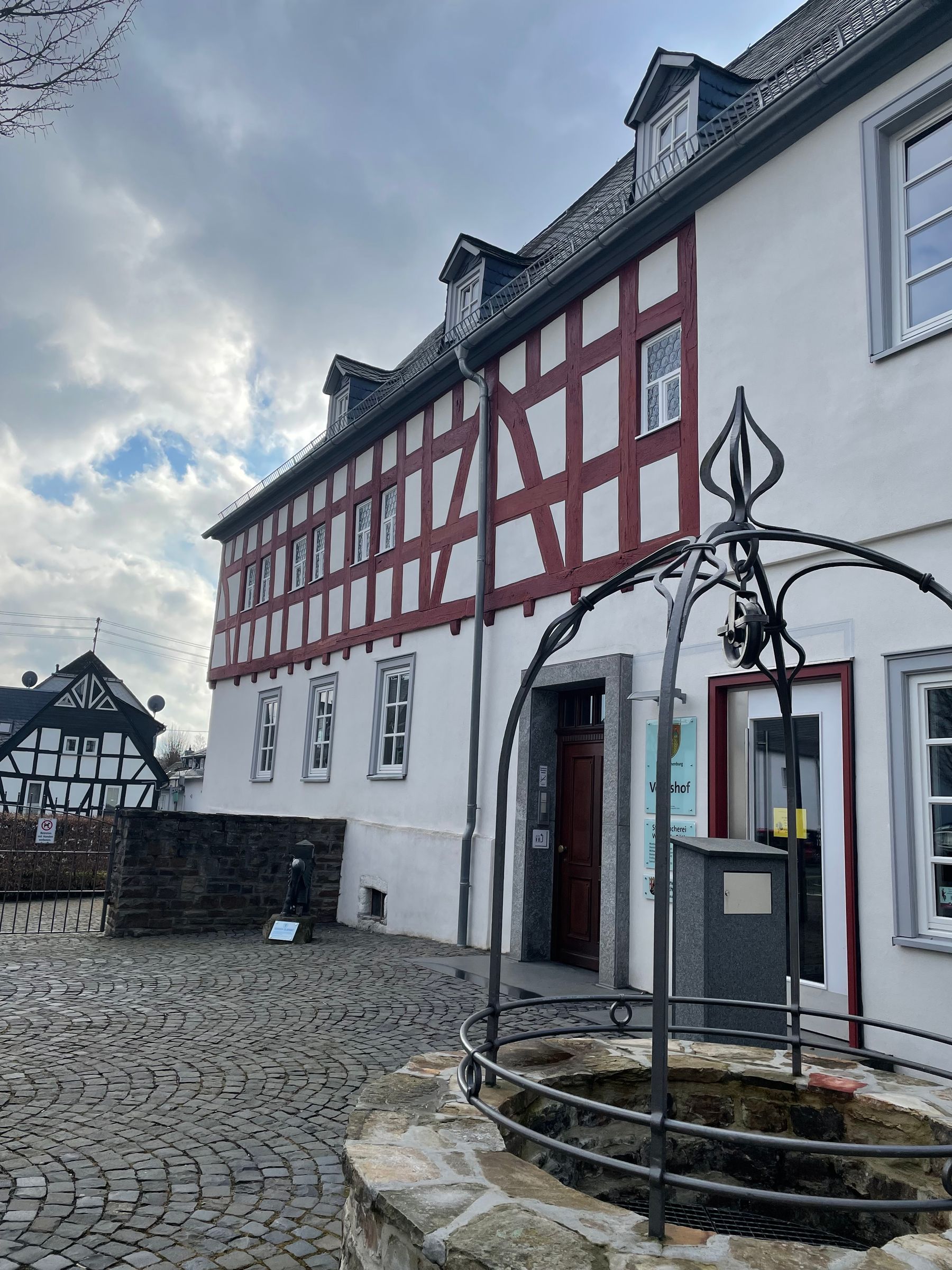 Ein traditionelles Fachwerkhaus mit weißen Wänden und roten Balken steht hinter einem steinernen Brunnen mit einer verzierten Metallabdeckung unter einem bewölkten Himmel. In der Nähe sind Kopfsteinpflaster und ein weiteres Fachwerkhaus zu sehen.