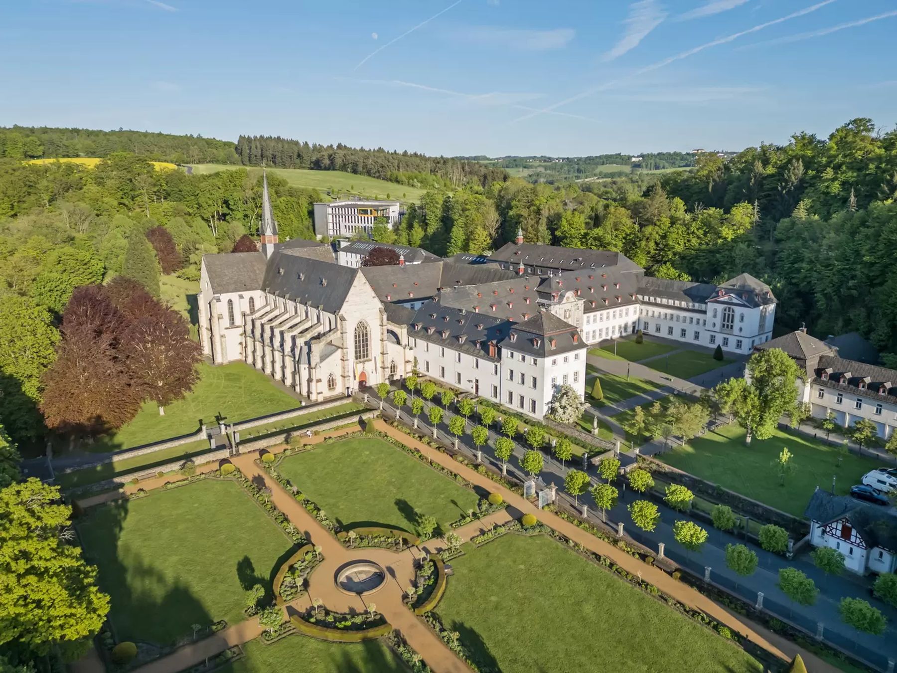 Luftaufnahme eines großen, historischen Klosterkomplexes mit weißen Gebäuden, einer Kirche mit hohem Turm, gepflegten Gärten und grünen Rasenflächen, umgeben von Bäumen und einer Landschaft unter blauem Himmel.