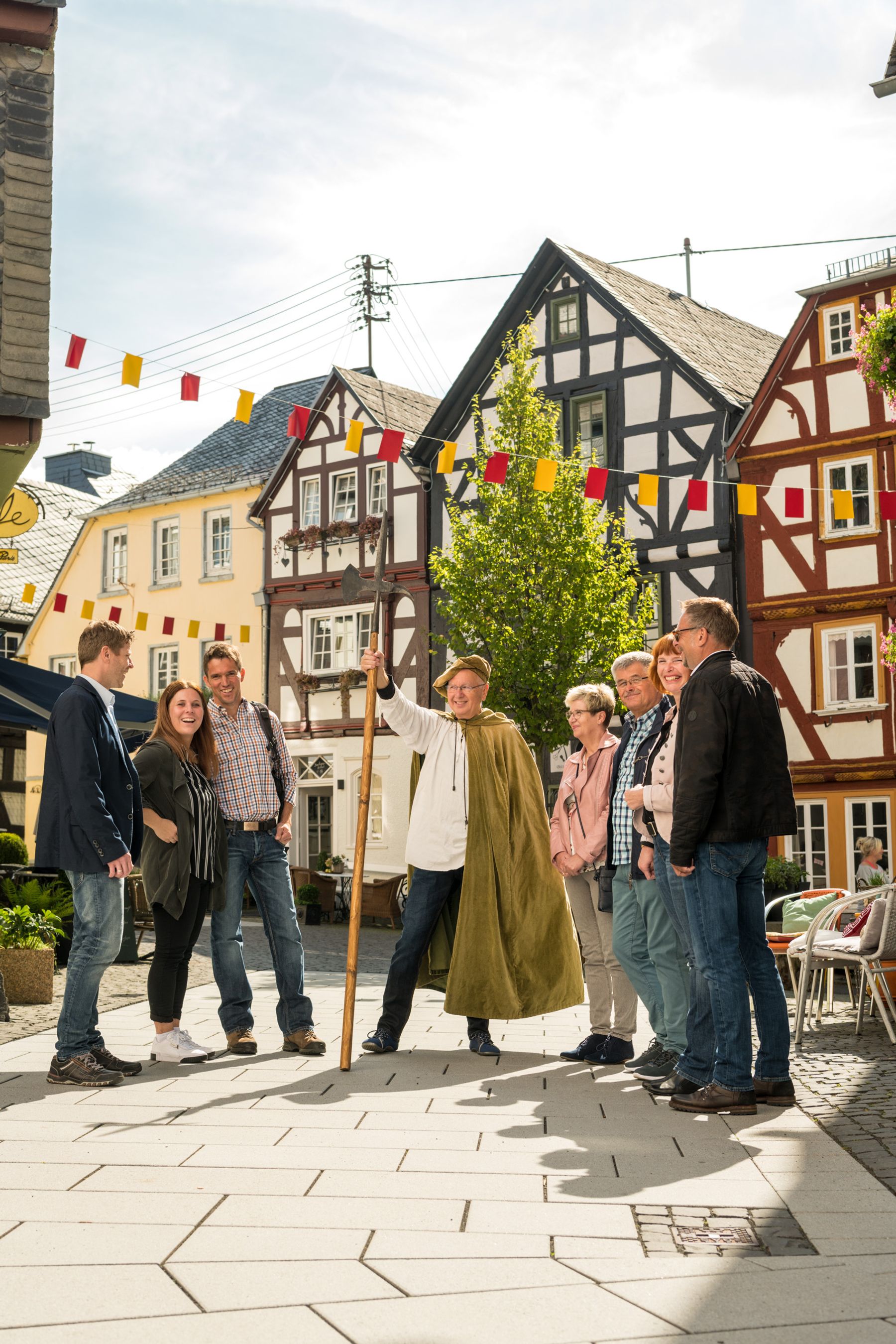 Eine Gruppe von Menschen steht und lächelt in einer sonnigen, alten europäischen Straße mit Fachwerkhäusern. Eine Person in der Mitte trägt einen Umhang und hält einen Stab, der die Gruppe zu führen oder zu unterhalten scheint. Bunte Fahnen hängen über ihnen.
