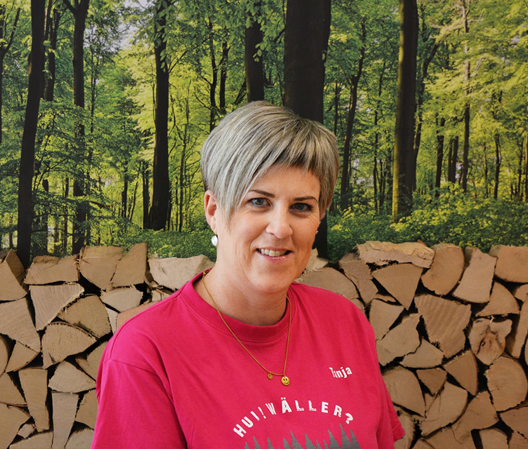Eine Frau mit kurzen grauen Haaren, einem rosafarbenen Hemd und einer goldenen Halskette steht vor einer Kulisse aus gestapeltem Brennholz und einer Waldszene.