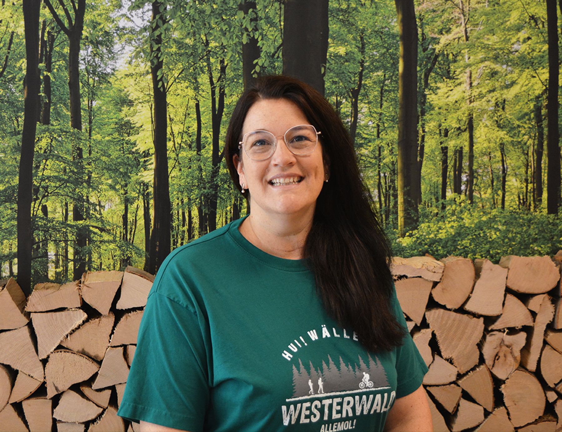 Eine Frau mit dunklem Haar und Brille lächelt in die Kamera. Sie trägt ein grünes "Westerwald"-T-Shirt und steht vor einer Kulisse aus gestapeltem Brennholz und einer Waldszene mit hohen Bäumen.