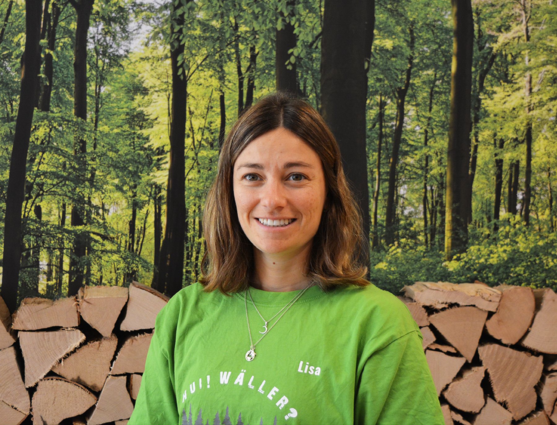 Eine Frau mit schulterlangen braunen Haaren, die ein grünes Hemd mit dem Namen Lisa trägt, steht lächelnd vor einer Kulisse aus gestapeltem Brennholz und einem üppigen grünen Wald.