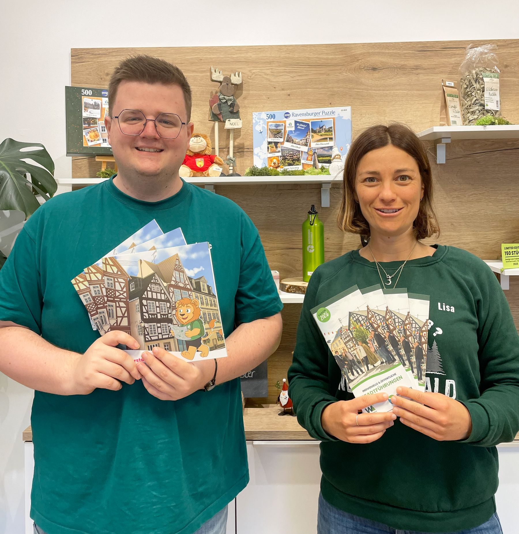 Zwei Personen stehen in einem Haus, lächeln in die Kamera und halten illustrierte Flugblätter oder Broschüren in der Hand. Sie stehen vor Regalen mit verschiedenen Souvenirs und Gegenständen, darunter Puzzles, eine grüne Wasserflasche und kleine dekorative Figuren.