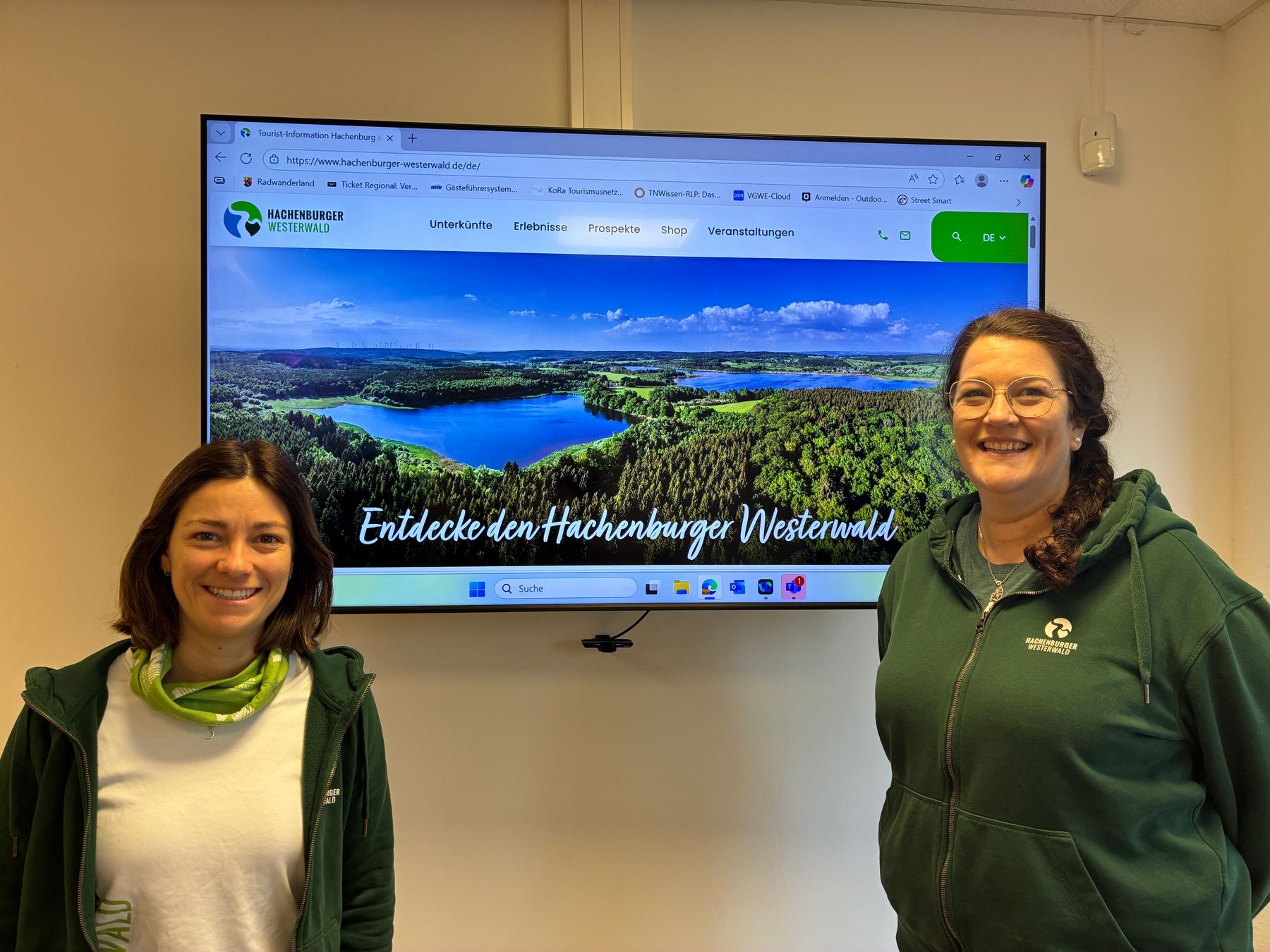 Zwei lächelnde Frauen in grünen Jacken stehen vor einem großen Bildschirm, der eine malerische Landschaft und die Website des Hachenburger Westerwalds in deutscher Sprache zeigt. Die Website zeigt einen Wald, einen See und die Worte "Entdecke den Hachenburger Westerwald".