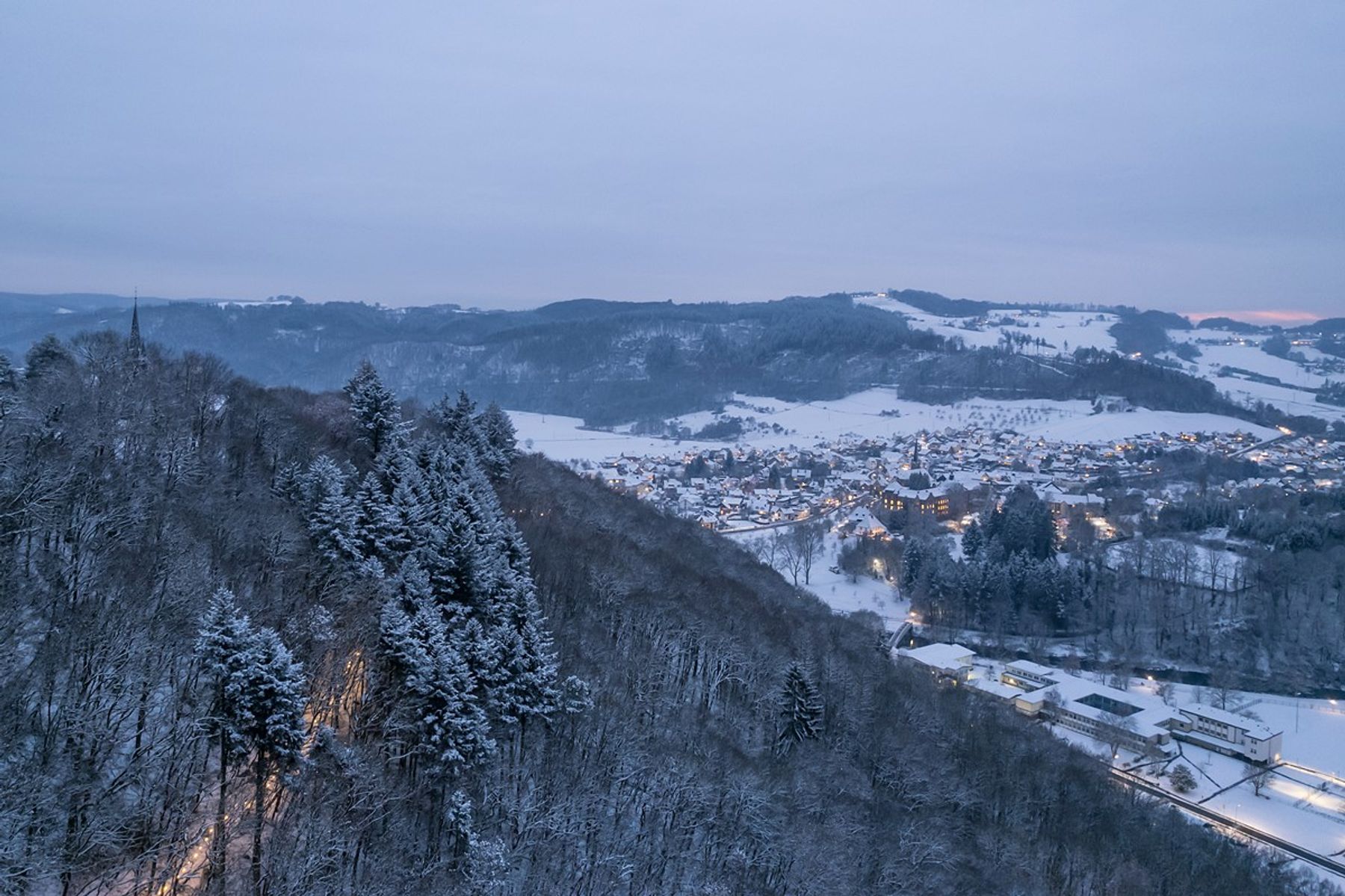 Schneebedeckte bewaldete Hügel überblicken eine kleine Stadt in einem Tal, deren Häuser und Gebäude von warmen Lichtern unter einem blassen, wolkenverhangenen Winterhimmel beleuchtet werden.